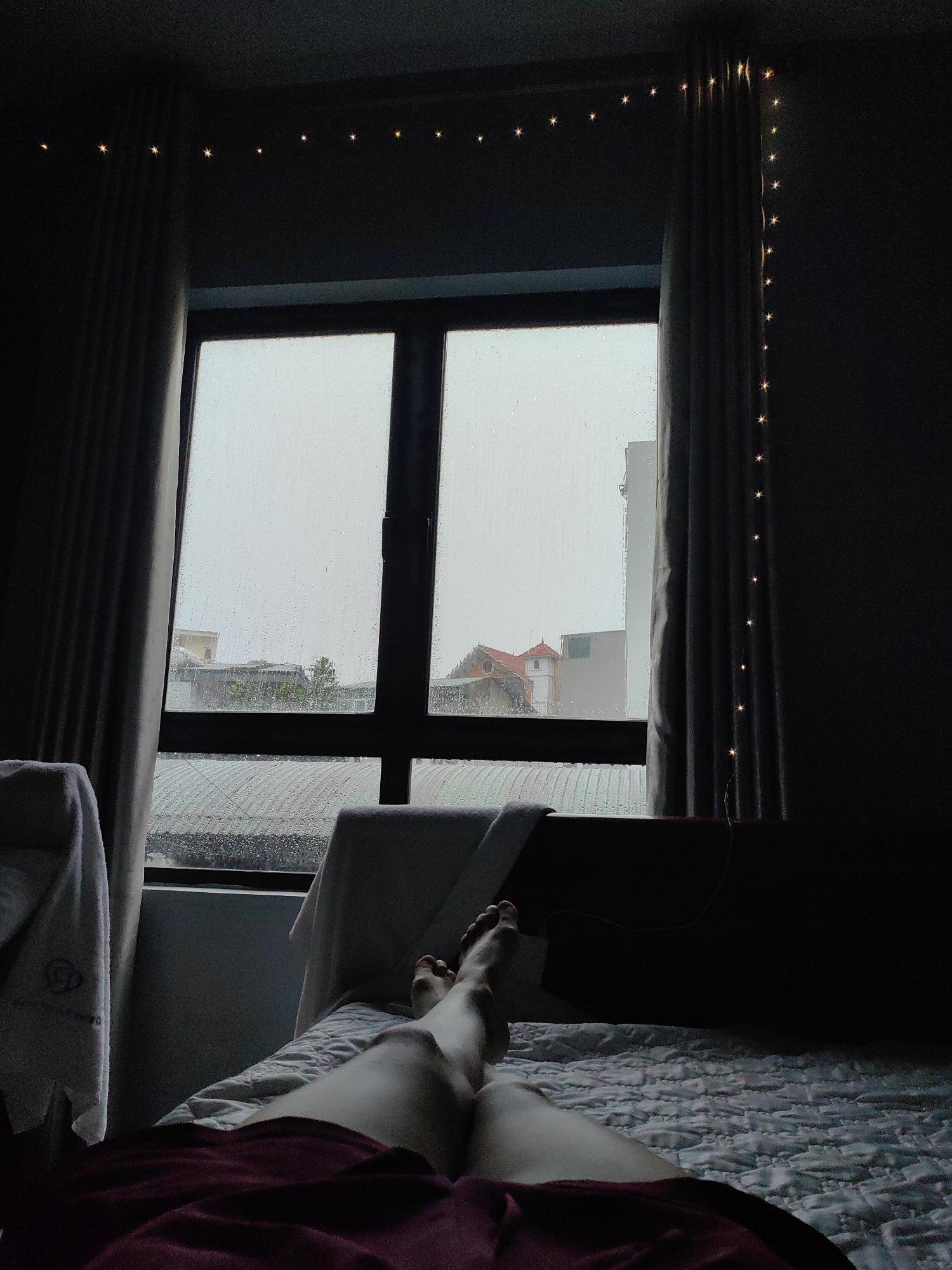 A person is lying in bed in a room. The window looks out onto a rainy day. Buildings are visible outside, characteristic of the urban setting. A string of lights decorates the room near the curtains. The location is likely Bai Chay Ward, Vietnam based on the architecture visible through the window.