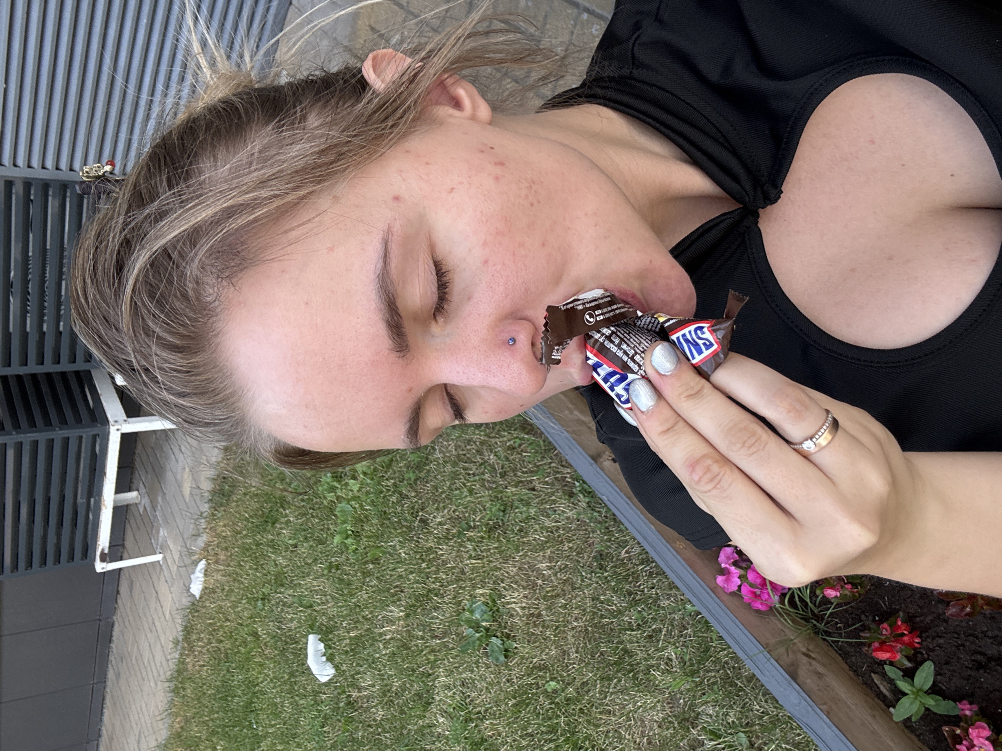 A young woman with blonde hair is eating a Snickers bar. She is outdoors, with grass and some pink and red flowers visible in a planter box. A building with grey horizontal siding and a white bench are in the background. The woman is wearing a black top with a cut-out detail at the chest. She has a nose ring and is wearing a ring on her left hand.
