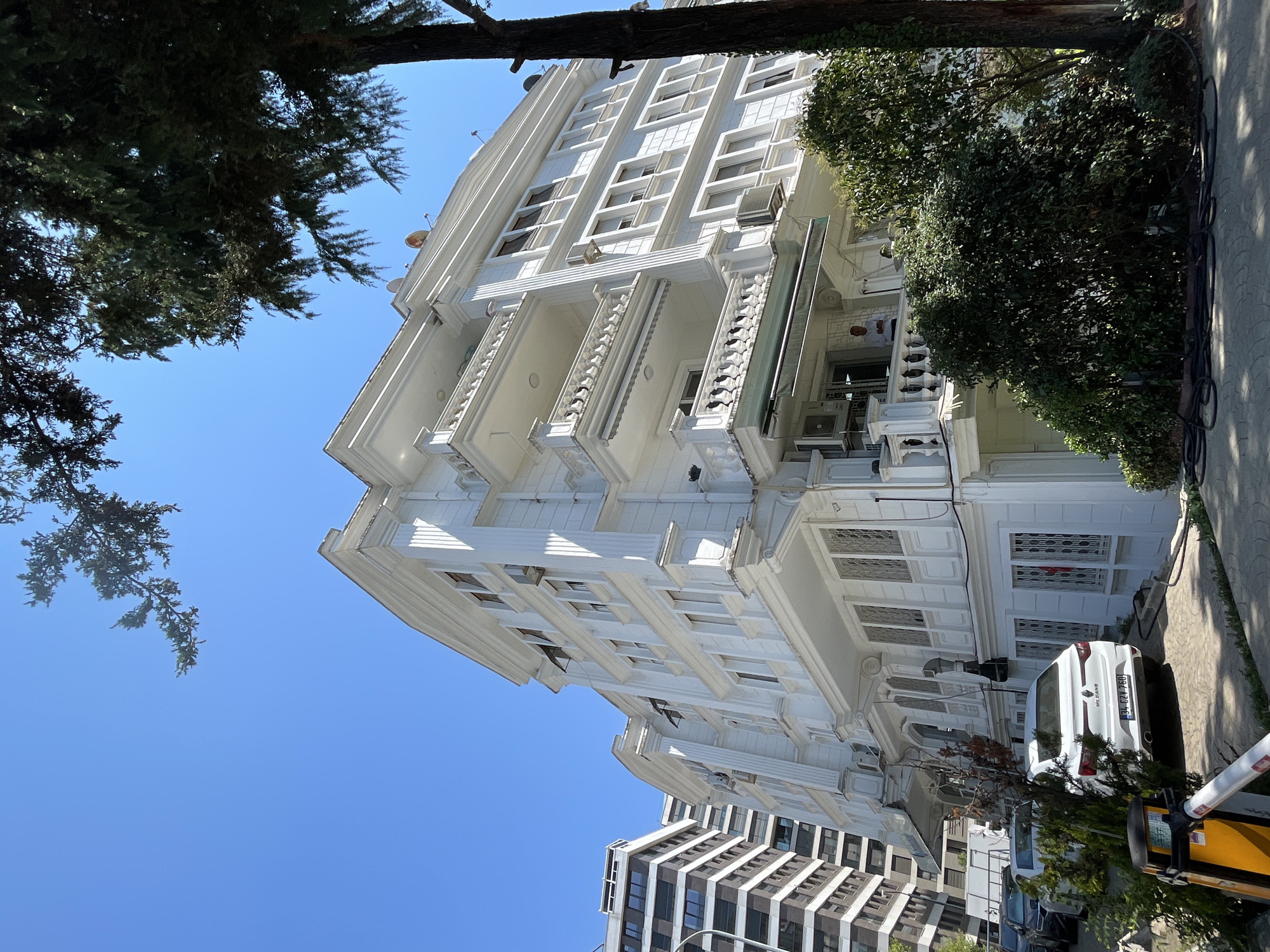 A white, multi-story building with ornate architectural details stands tall under a clear blue sky. The building features multiple balconies with decorative railings and numerous windows. Trees with dark green foliage are visible in the foreground and partially obscure the building. To the left, a modern, taller building with a dark facade and white horizontal stripes suggests an urban setting. Several cars are parked in front of the white building, including a white sedan with a license plate reading "34 CZV 760." A yellow and black barrier gate is also present in the foreground. A man in a light-colored shirt can be seen standing on one of the balconies. The overall scene depicts a residential or commercial area in the city of Kadıköy, Turkey, on a sunny day.
