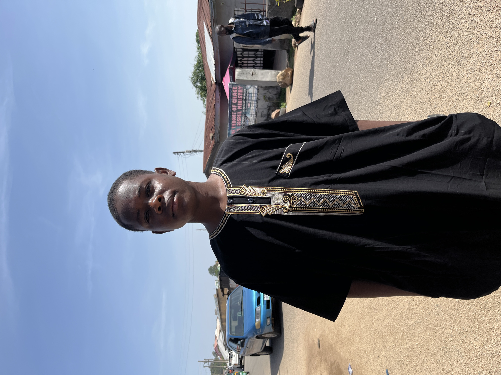 A young Black man is prominently featured, standing outdoors and looking directly at the camera with a neutral expression. He is wearing a black, long-sleeved traditional tunic, likely a kaftan or agbada, adorned with elaborate gold and silver embroidery around the V-neckline and down the chest.
The scene is an urban or semi-urban street, bathed in bright daylight under a clear blue sky with a few wispy clouds, suggesting midday or early afternoon. The ground is a paved road, possibly asphalt, with some dirt or sand visible on the sides.
In the background, on the viewer's left side of the street, there are several buildings. One building has a rusty corrugated iron roof and a prominent sign that reads "THE THRONE OF GRACE GOSPEL MINISTRY" with smaller text below, including "CHURCH" and "SCHOOL OF THE WORD". Further down the street, another person, dressed in a denim jacket and dark trousers, is walking away from the camera. A blue car, possibly a van or small bus, is also visible on the street behind the main subject, along with other indistinct vehicles further in the distance. The overall setting appears to be a typical street scene in Jos, Nigeria. The image is originally rotated 90 degrees clockwise.