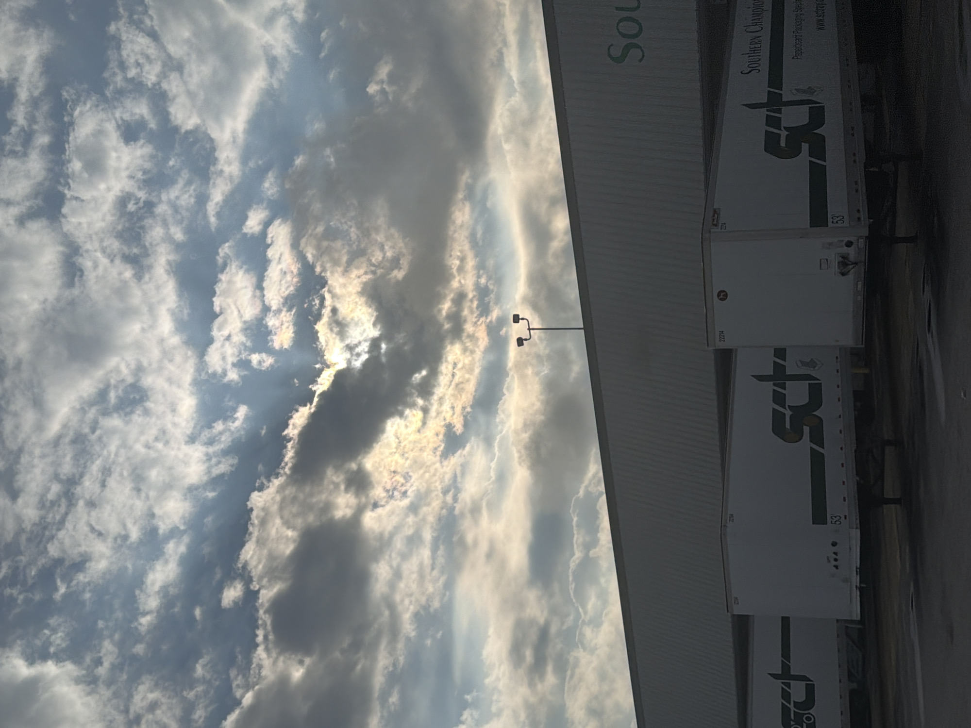 A bright, iridescent cloud formation with the sun peeking through is visible in a grey and blue sky above a long, low-rise industrial building. The building has a light-colored facade, and a sign with the word "Sou" is partially visible on the right side. In front of the building, several white semi-trailer trucks are parked. The trucks have green lettering that appears to be a logo and the words "SOUTHERN CHAMPION". The scene is likely an industrial area or warehouse in Chattanooga, United States of America.