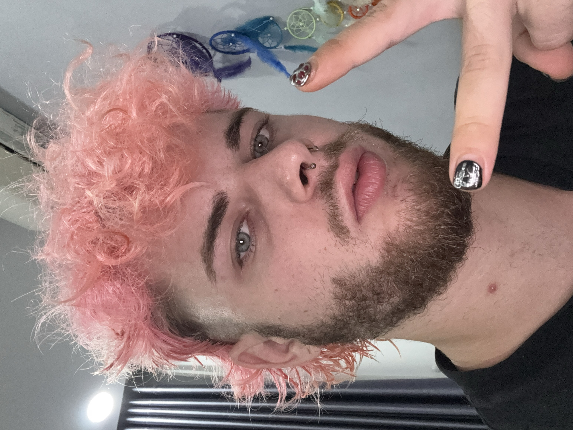 A close-up, rotated head-and-shoulders portrait of a young man with light skin. He has a beard, a piercing on his left nostril, and short pink curly hair. He is wearing a black shirt. His fingernails are painted, one with a silver design, the other with a red-and-silver design. In the blurred background above his head are several dreamcatchers. The location is not discernable from the image.