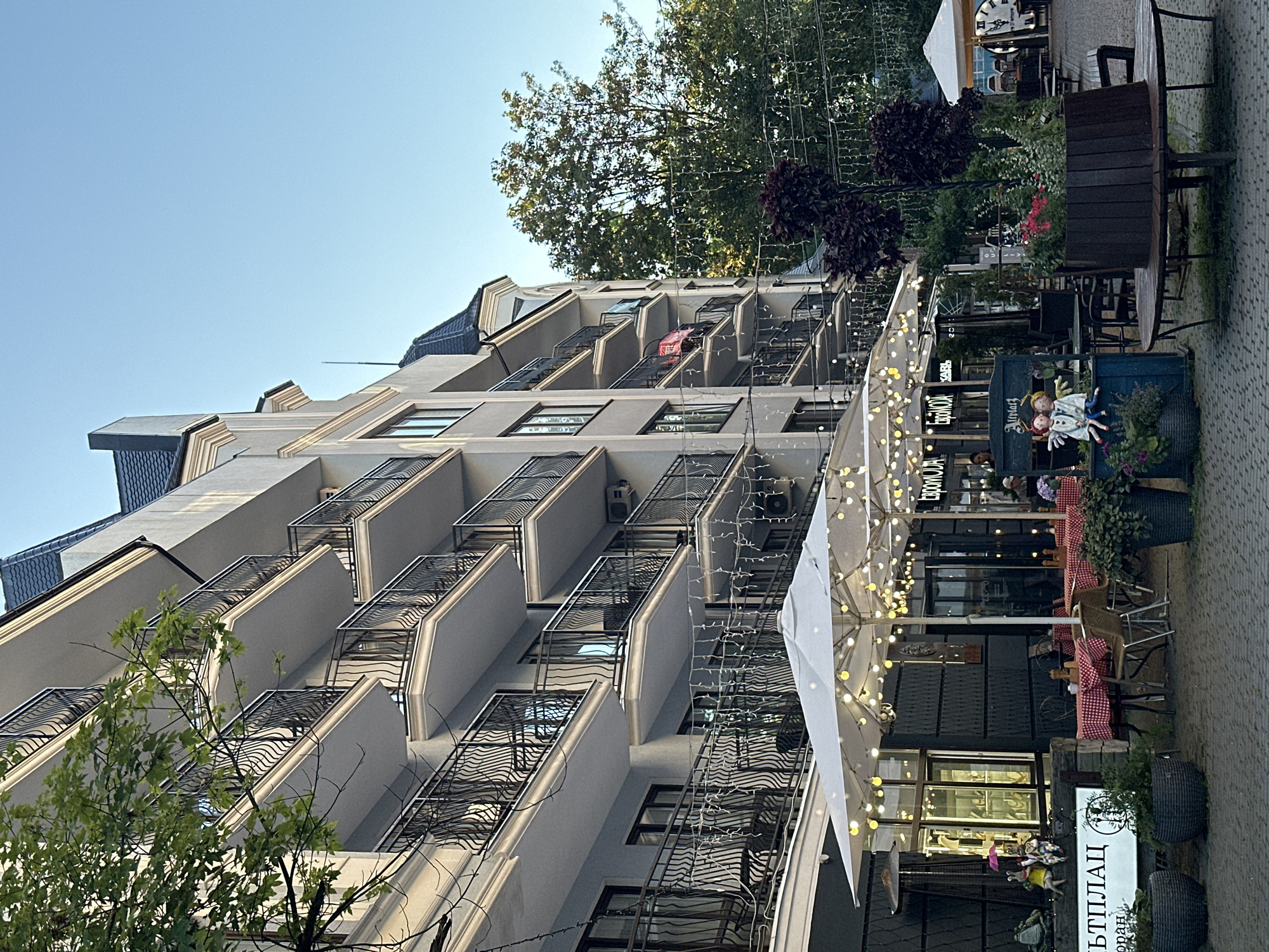 The image shows the exterior of a multi-story building in Svetlogorsk, Russia, with balconies and a ground-level outdoor seating area for a cafe or restaurant. The building's facade is light-colored with dark railings on the balconies. The outdoor seating area is set up with tables covered in red and white checkered tablecloths, chairs, and large white umbrellas adorned with string lights. In front of the seating area, there are potted plants and decorative figures. Beyond the cafe, there are more trees and other structures visible. The sky is clear and blue.