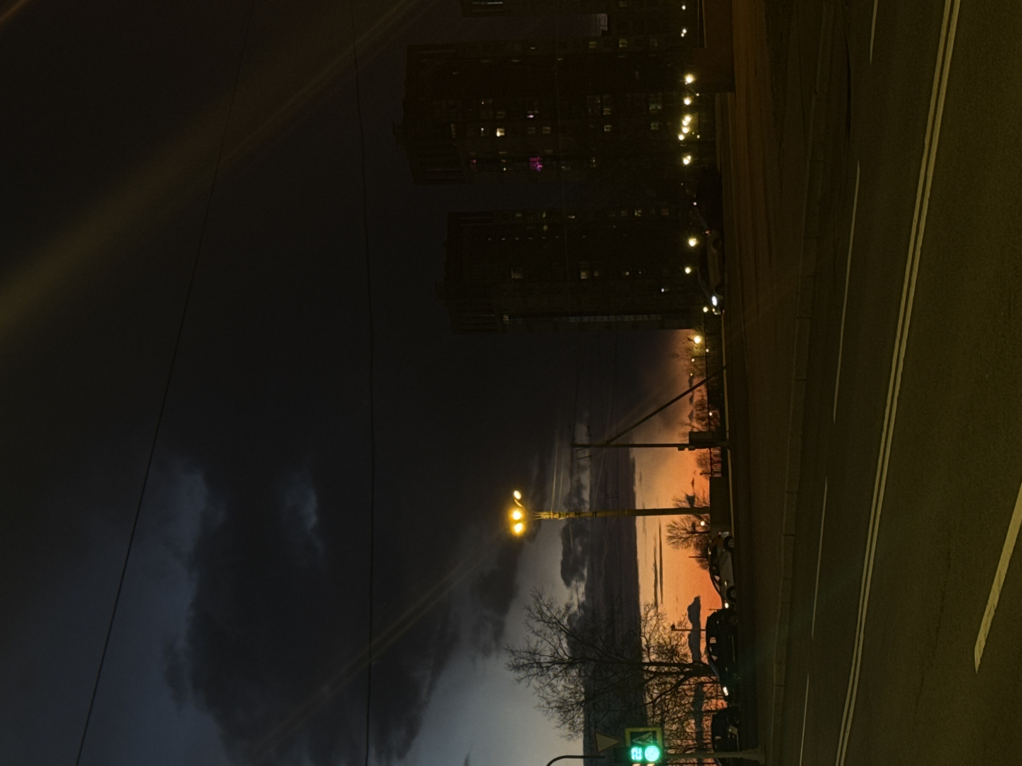 This media file captures an urban scene during twilight, likely dusk or dawn, in Saint Petersburg, Russia. The image is oriented vertically but depicts a horizontal landscape.
The sky is a prominent feature, transitioning from dark, heavy grey-blue clouds in the upper-left to a striking orange, red, and yellow horizon on the right. Silhouettes of bare tree branches are visible against this colorful backdrop, suggesting a late autumn or winter season.
A multi-lane road occupies the foreground and right side of the image, marked with clear double white lines separating traffic lanes and a single white line on the shoulder. Streetlights are illuminated, casting yellow light across the scene; one prominent streetlight pole stands in the mid-ground. In the lower-right corner, a green traffic light is visible, displaying the digital number "23" in white, possibly indicating a countdown. A yellow triangular road sign is partially obscured below the traffic light.
In the background, dark, multi-story buildings with numerous illuminated windows, some with a purple or pink hue, rise against the dim sky. Other distant lights punctuate the darkness. A few dark car shapes are discernible on or near the road, and overhead wires traverse the sky. The scene is quiet, devoid of prominent human activity, capturing a serene moment in a city at the cusp of night or day.