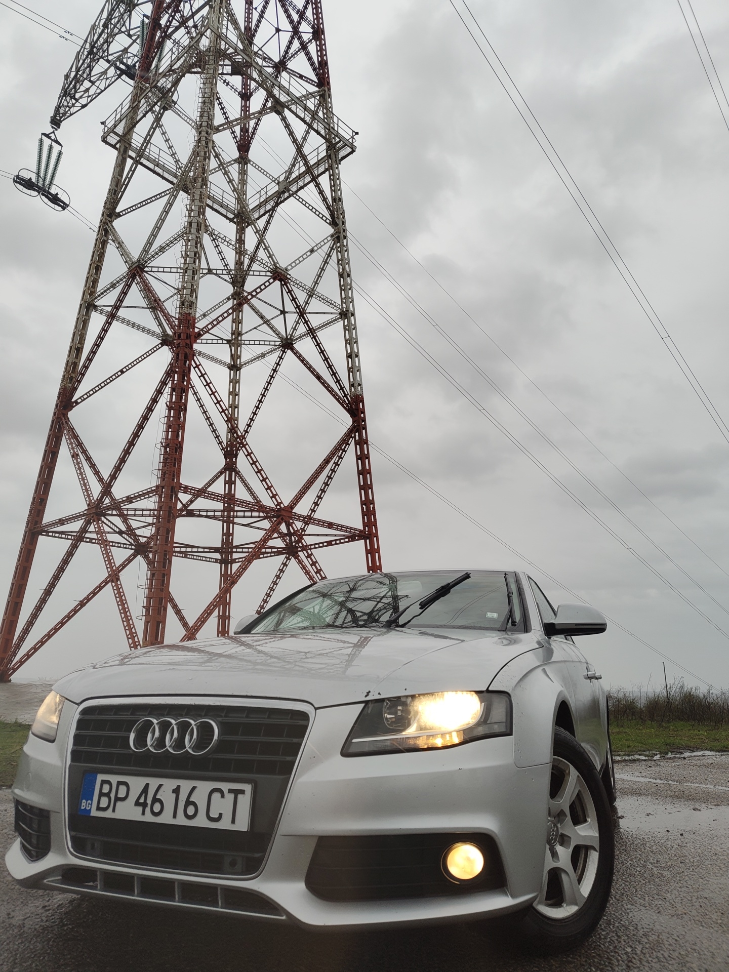 A silver Audi sedan, bearing Bulgarian license plate 'BP 4616 CT', is positioned on a wet, unpaved surface in Kozloduy, Bulgaria. The front of the vehicle is angled slightly left, with its headlights and right fog light illuminated. The windshield wipers are in a raised position. Dominating the background is a large, lattice-steel electricity transmission tower, featuring red-brown and grey structural elements, from which high-voltage lines extend across an overcast, grey sky. Sparse vegetation lines the distant horizon to the right. No individuals are visible in the scene.