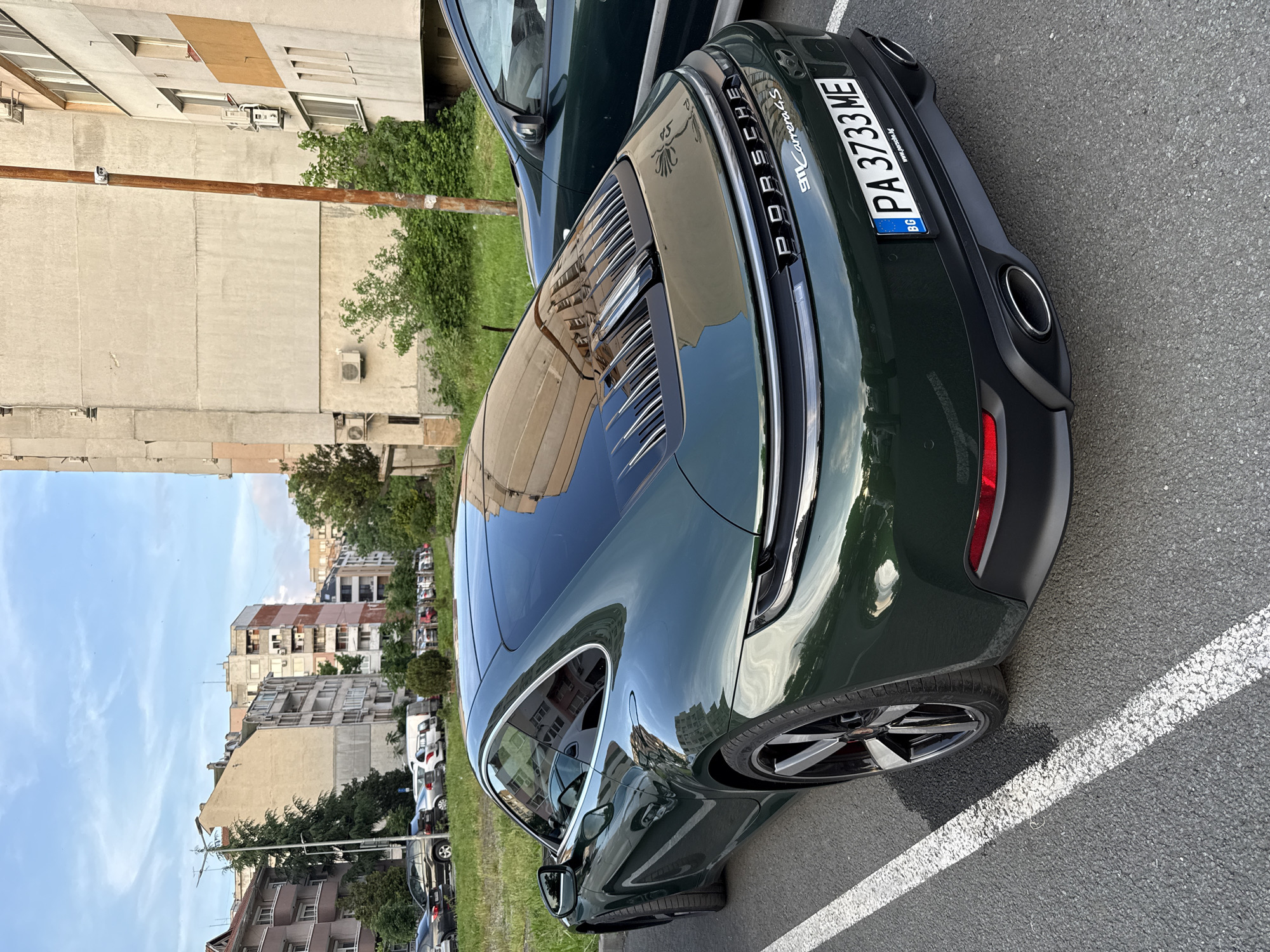 The image shows the rear of a dark green Porsche 911 Carrera 4S parked on asphalt with a white parking line next to the rear tire. The license plate reads "PA 3733 ME" and indicates Bulgaria (BG). The car has the "911 Carrera 4S" emblem. In the background, there are buildings characteristic of an urban setting, suggesting the location is in the city of Burgas, Bulgaria. A utility pole is visible in the mid-ground, along with trees and other parked vehicles. The buildings are of varying heights, and their architectural styles appear typical of residential areas in the city. The sky is visible between the buildings and is a light blue color with some clouds.