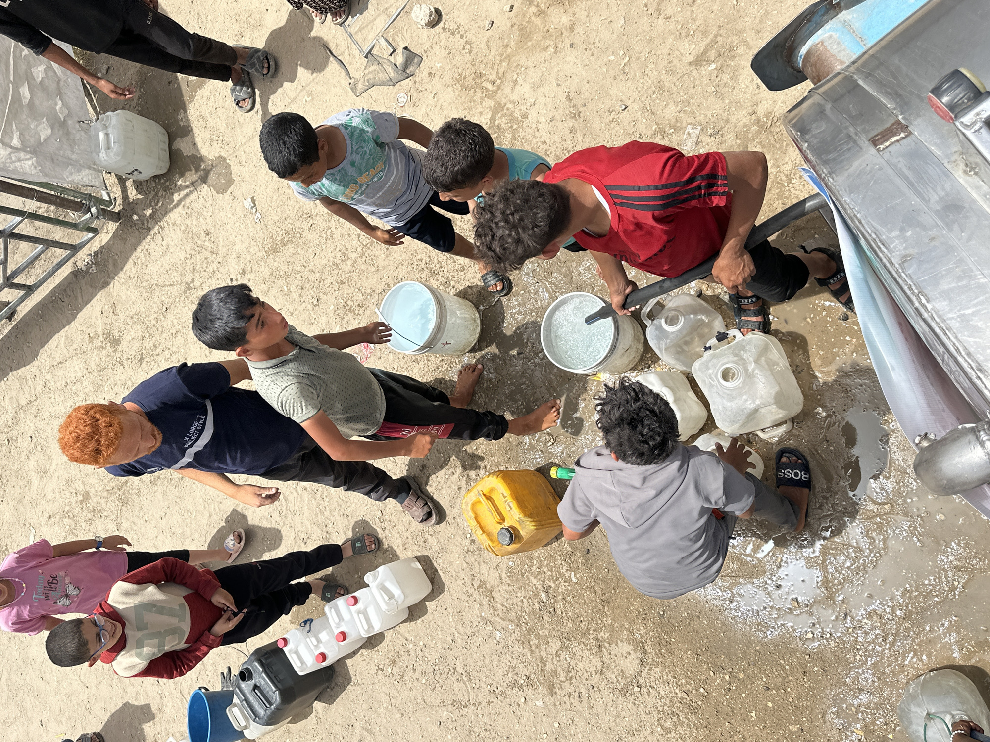 In Gaza City, Palestinian Territories, a group of children are filling various containers with water from a truck. The containers include white plastic jugs, buckets, and a yellow jerrycan. The ground is wet and sandy. The children are wearing casual clothing, and some are barefoot or wearing sandals. The scene appears to be in a residential area with some kind of metal structure in the background.
