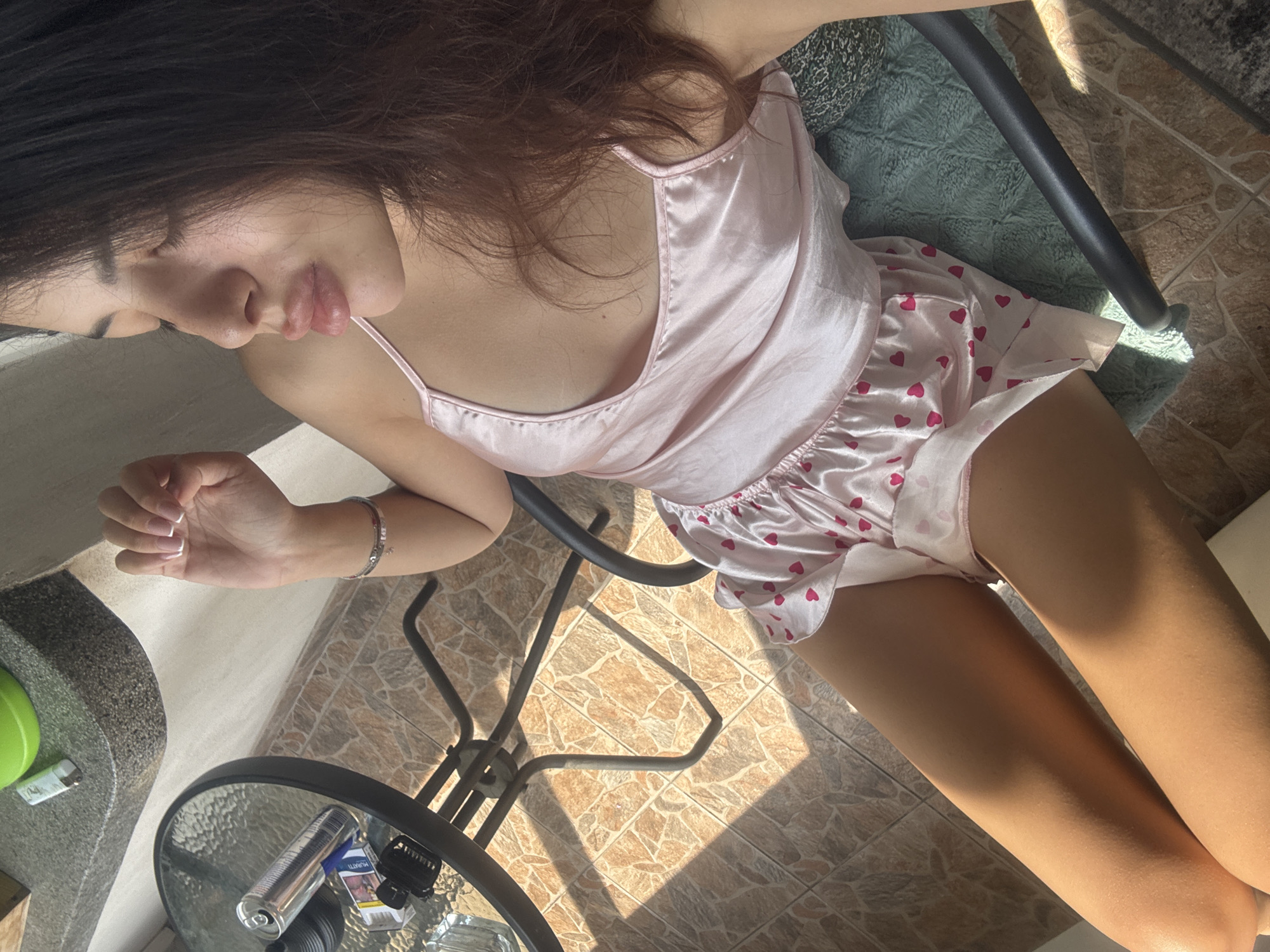 A young woman with long, dark hair is seated outdoors on a chair, her body angled towards the right. She is wearing a light pink satin camisole with thin straps and matching pink satin shorts adorned with small red hearts. Her legs are crossed, and she is looking downwards, her lips slightly parted. Sunlight casts shadows from a nearby table and chair onto the tiled floor. On the table, to the left, are a can, a pack of cigarettes, and a lighter. The setting appears to be a patio or balcony with stone tiles on the floor. This image does not contain enough information to determine its location within the city of Plovdiv, Bulgaria.