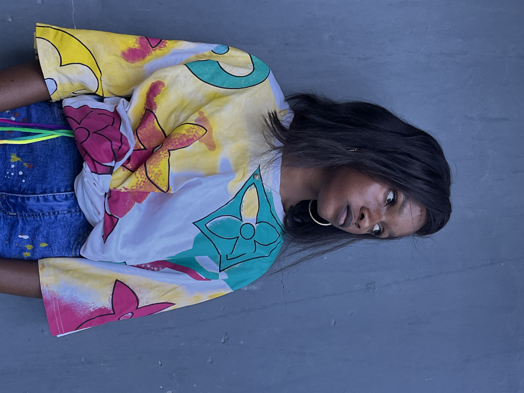 A person is lying on a gray surface outdoors. The person has dark hair and skin, and is wearing gold hoop earrings. The person is wearing a colorful shirt with floral patterns in yellow, pink, green, and white, along with denim bottoms that have white paint splatters and colorful cords. The background consists of a plain gray surface. The location or city is not possible to determine based on only the image provided.
