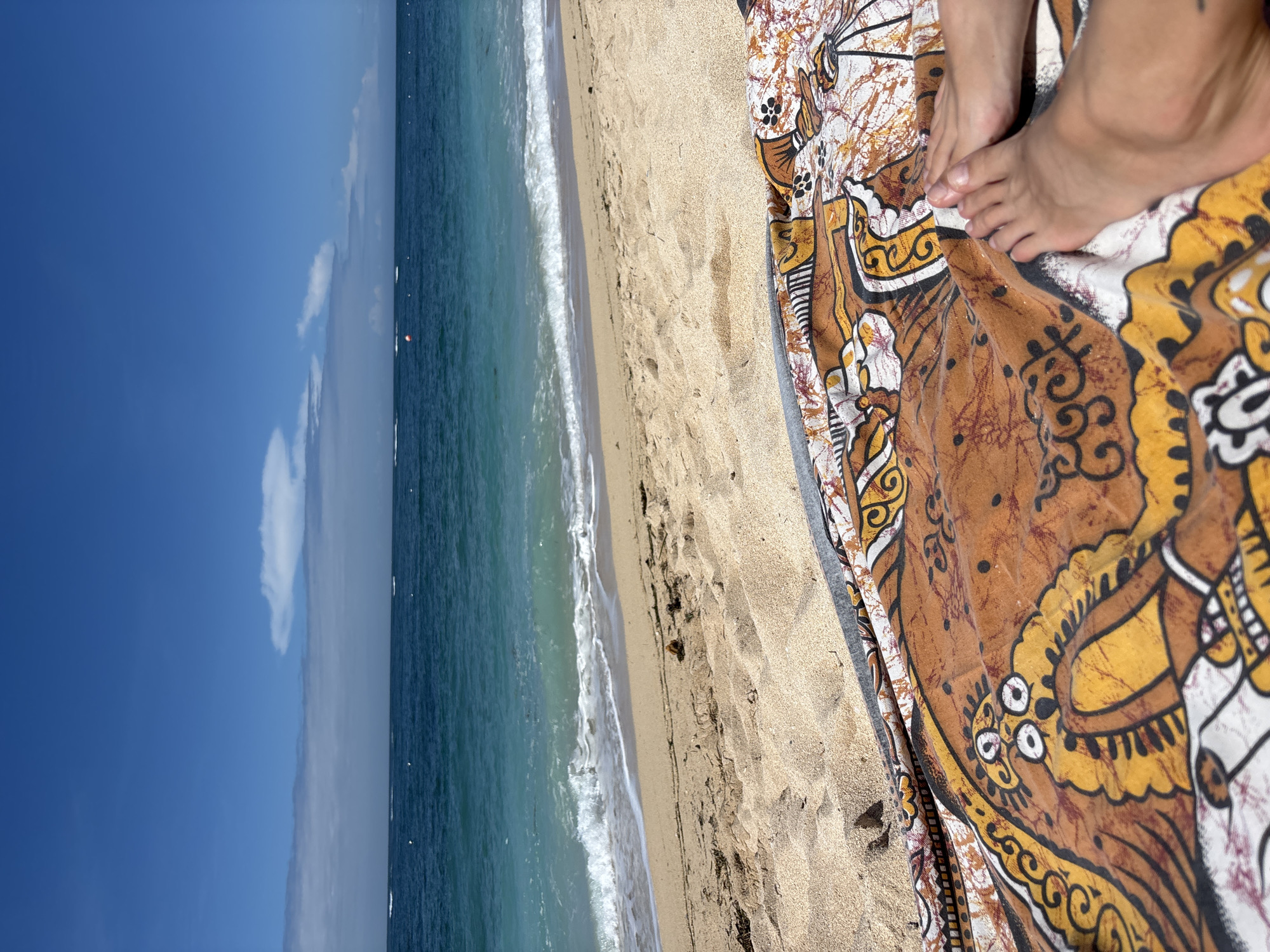 The image depicts a beach scene, possibly in Kuta Selatan, Indonesia. The view includes a turquoise ocean with small waves meeting a sandy beach. A patterned brown and white cloth lies on the sand with a person's feet resting on it. The sky is blue with a single white cloud.