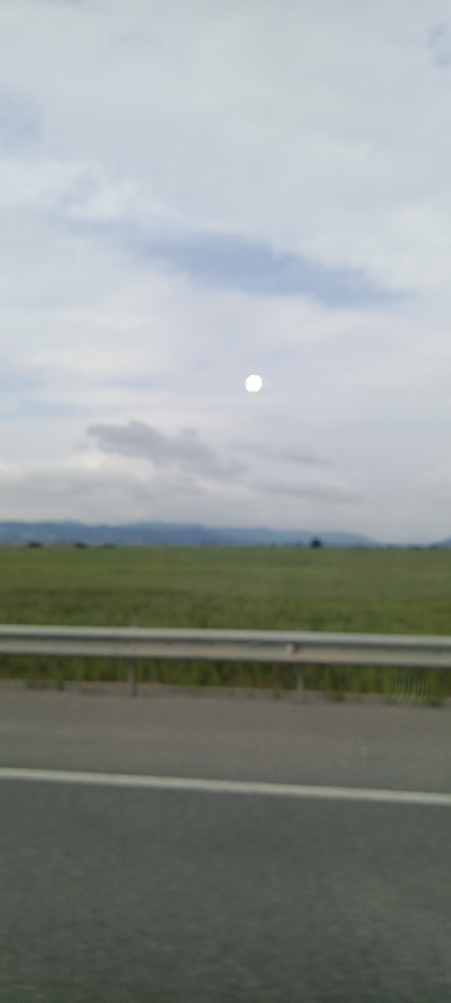A bright, round object, possibly the sun or moon, is visible in a cloudy sky over a rural landscape. In the foreground, there is a road with a white line and a metal guardrail. Beyond the guardrail, a vast green field stretches to the horizon, where distant, hazy mountains can be seen. The image appears to have been taken from a moving vehicle. There are no people or animals visible in the frame. The overall scene is overcast and somewhat blurry, suggesting it may be a photograph taken with a smartphone or a low-quality camera. The location is identified as Shkotovo, Russia, although no specific landmarks or indicators of this location are present in the image itself.