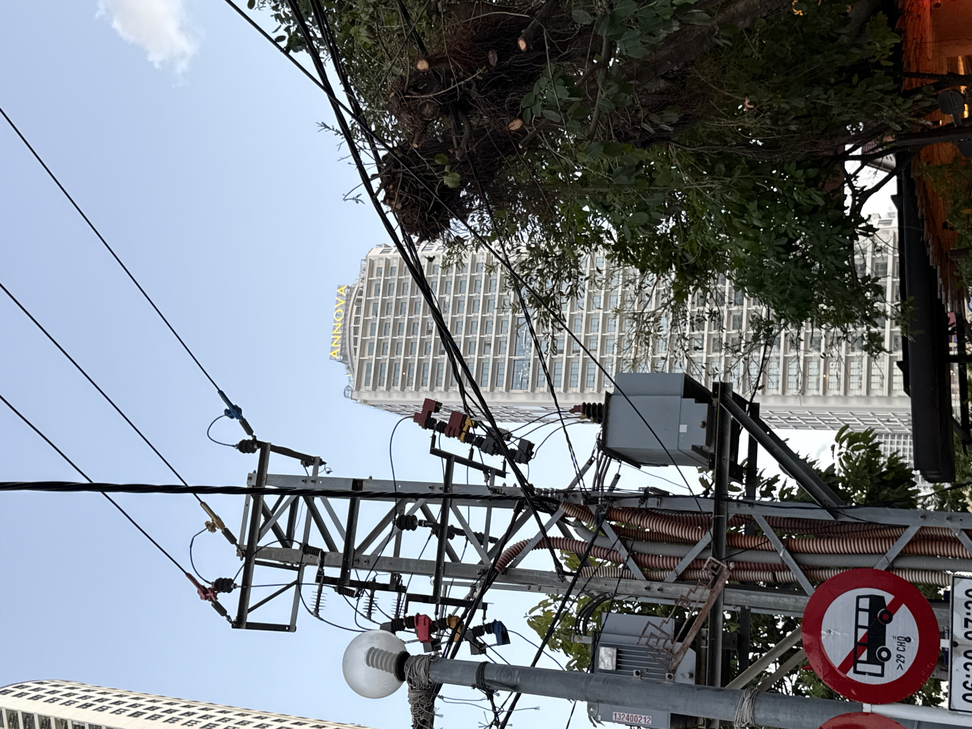 The image depicts an urban scene in Nha Trang, Vietnam, characterized by a complex network of electrical poles, wires, and a prominent high-rise building. In the foreground, a utility pole is laden with various electrical components, including transformers, insulators, and a street lamp. Several wires crisscross the frame, obscuring parts of the sky and the background. A circular red and white traffic sign prohibiting buses carrying more than 29 passengers is visible on the left side of the pole.
Behind the electrical infrastructure, a tall, modern building with numerous windows and a distinctive illuminated sign that reads "ANNOVA" stands out against the clear blue sky. The building's facade suggests a commercial or residential complex. To the right of the building, a large tree with dense foliage and branches intertwines with some of the electrical wires, creating a visual overlap between nature and urban infrastructure. The overall impression is one of a busy, developing city where technology and nature coexist, often in close proximity. The lighting suggests it is daytime, possibly late afternoon given the slight warmth in the sky.