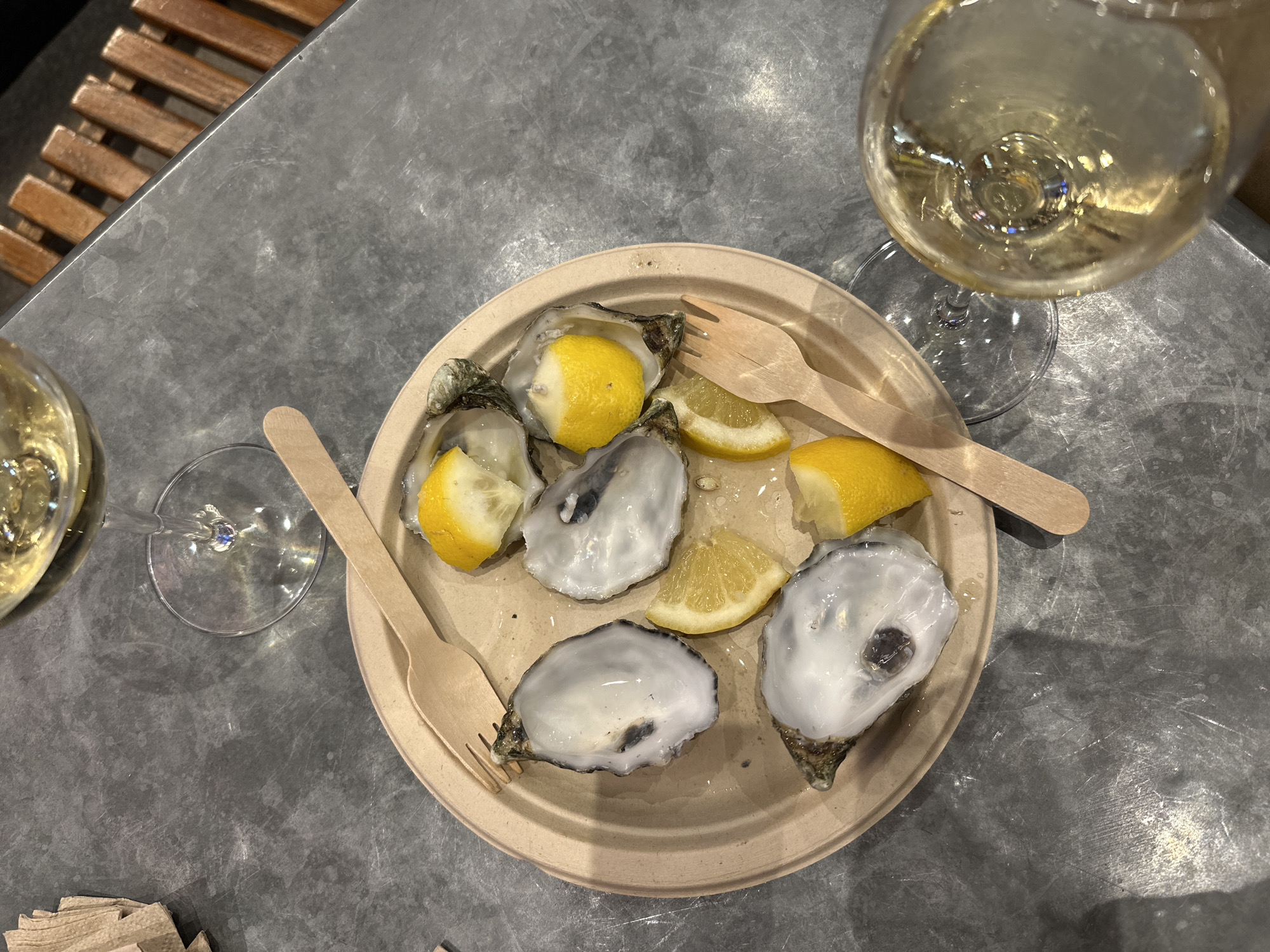 The image shows a tabletop scene, possibly in a restaurant or bar in Madrid, Spain. The focus is on a round, light-beige disposable plate holding four opened oysters, each topped with a slice of lemon. The plate also contains a couple of additional lemon slices. Two light-brown wooden forks rest on the plate. To the upper right and left, two glasses of white wine are visible. The table surface appears to be metallic and textured, with a visible grain. At the extreme left edge, a glimpse of a wooden structure (possibly a chair or bench) can be seen. In the bottom left corner, there's a stack of napkins. The scene is illuminated, with overhead lighting casting shadows and highlights.
