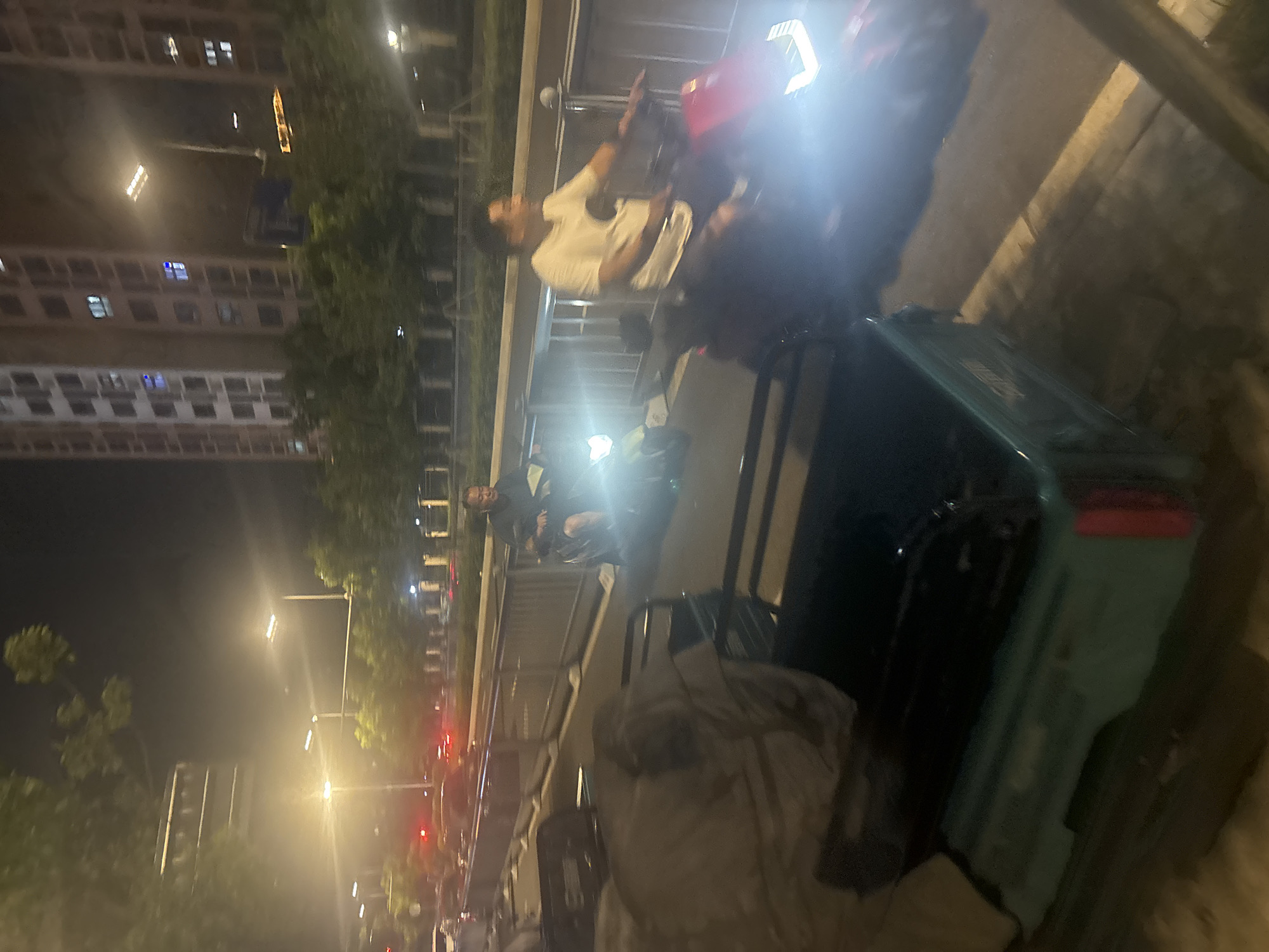 In Suzhou Industrial Park, China, at night, two men are riding electric scooters on a street. The man on the left is wearing a dark shirt and the man on the right is wearing a white t-shirt. They are riding side-by-side, with the man on the right looking to his left and smiling. The scooters have bright headlights that illuminate the road. In the foreground, there is a teal-colored electric tricycle with a black cargo box. In the background, there are tall buildings, trees, and streetlights. The scene suggests a typical urban evening in a developing area.