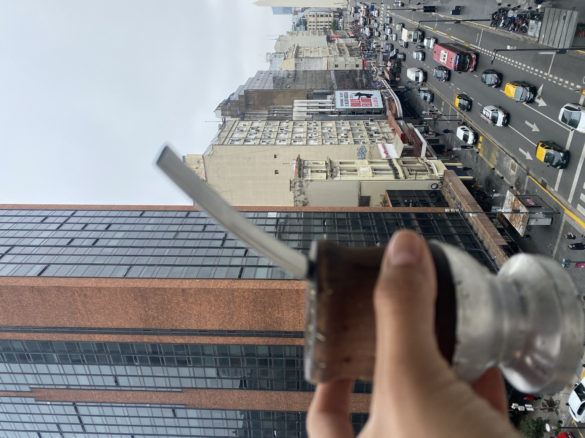 A slightly angled eye-level shot depicts a person holding a mate in the foreground, with a cityscape of Buenos Aires, Argentina in the background. The mate is made of wood and glass with a metal straw. The building in the foreground is a modern high-rise with glass and brick accents. The background reveals a bustling city street lined with various buildings, including older architectural styles. Heavy traffic includes cars, taxis, and a red bus. Pedestrians are visible on the sidewalks. The Obelisco, a prominent landmark, can be seen in the distance. The sky is overcast.