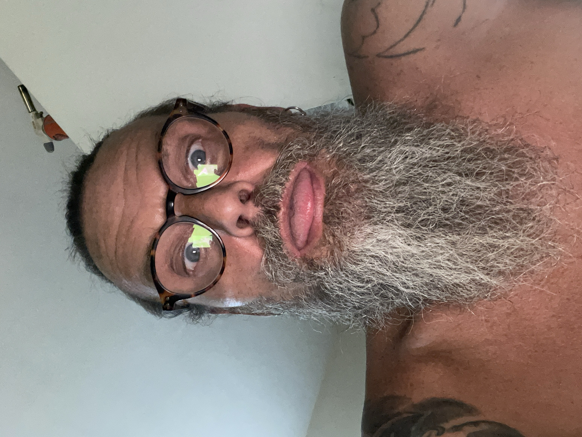 A close-up selfie shows a shirtless man with a grey beard, glasses, and his tongue sticking out. The background is a plain wall. The lighting suggests an indoor setting. There is a small orange and silver object attached to the wall in the upper right portion of the image. This object appears to be a small torch or a similar handheld device.