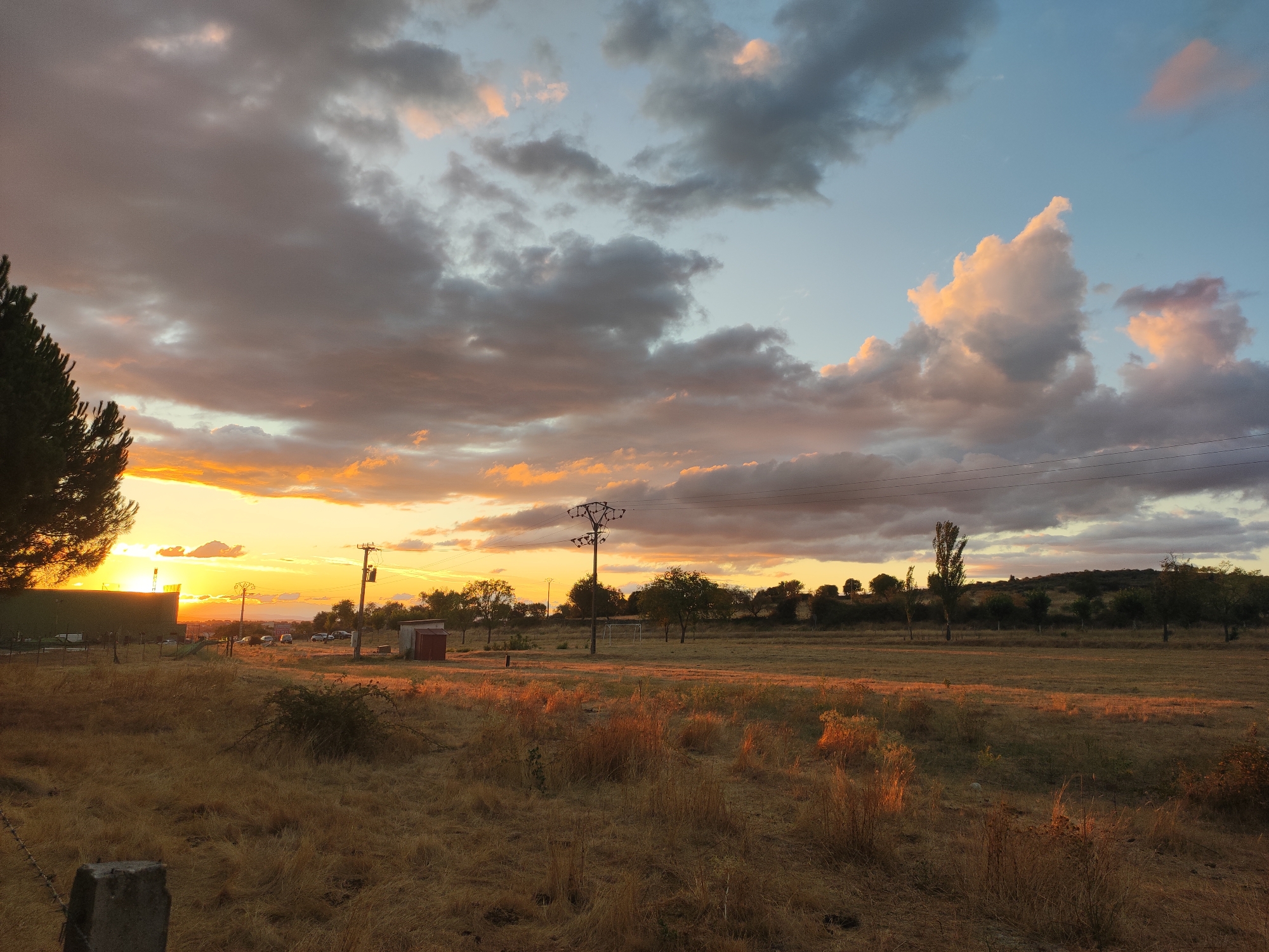The image captures a sunset over a rural landscape in the vicinity of Sobradillo, Spain. The sky is a dramatic canvas of warm oranges, yellows, and blues, streaked with gray and pink clouds. In the foreground, dry, golden grass covers a field, with a concrete post and barbed wire visible on the left, suggesting agricultural land. A dark green building or structure is partially visible on the far left, silhouetted against the bright sunset. Further into the scene, a dirt road leads towards a cluster of parked vehicles. A tall electricity pylon stands prominently in the middle ground, with a small red shed to its left. A sparse line of trees and undulating hills form the horizon under the colorful sky. The overall impression is one of a serene, late afternoon rural setting.