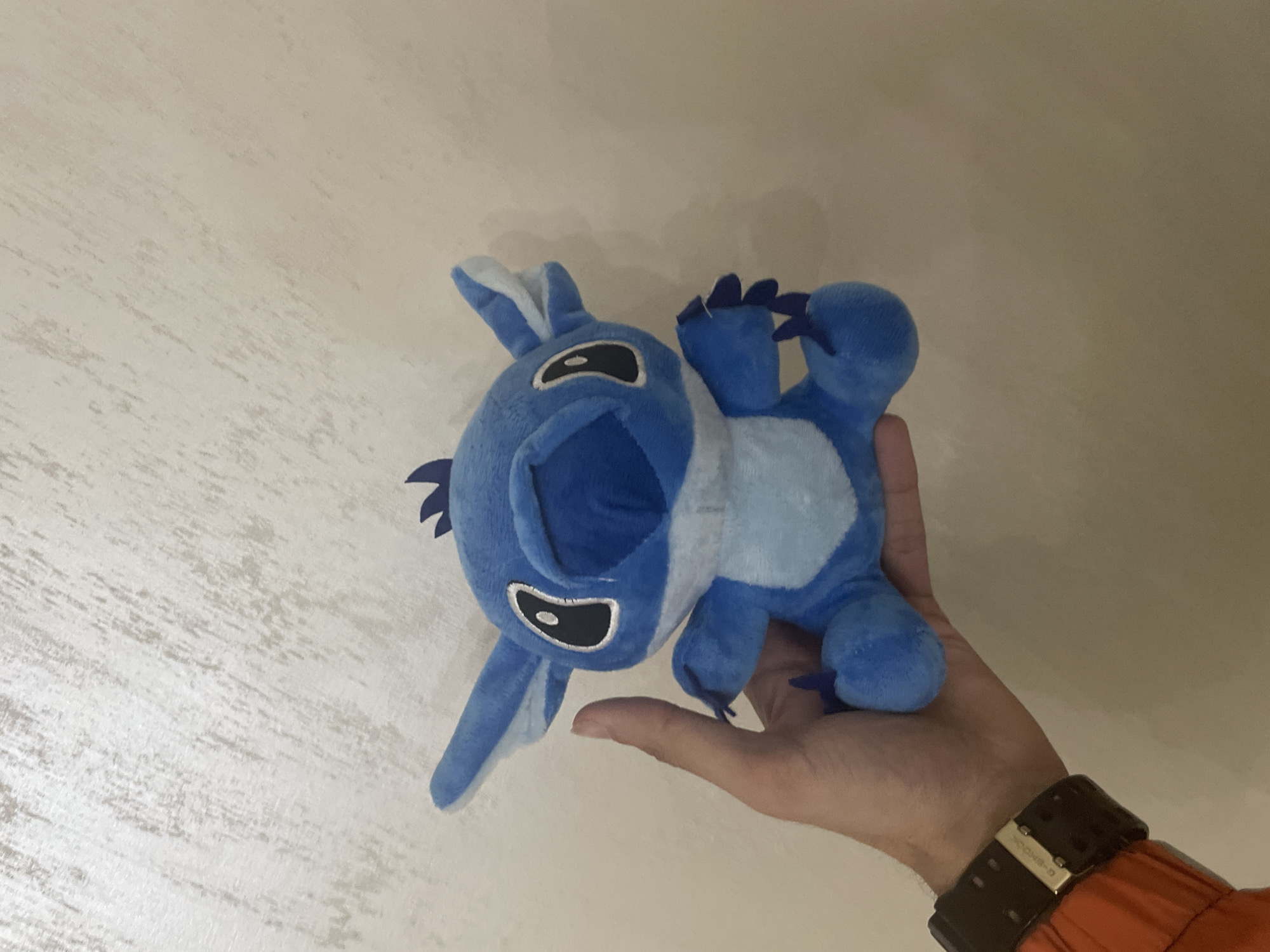 An individual's light-skinned hand, positioned on the left side of the frame, holds a blue Stitch plush toy against a light beige textured wall in Ihtiman, Bulgaria. The plush, predominantly blue with lighter blue sections on its chest, inner ears, and around its mouth, features black oval eyes with white pupils and black outlines. Small, dark blue spikes are present on the top of its head and on its feet. The hand wears a black G-Shock digital wristwatch with a silver buckle on the left wrist. An orange garment, consistent with a sleeve, is visible on the lower left quadrant beneath the watch. The wall in the background exhibits a subtle, irregular textured pattern.