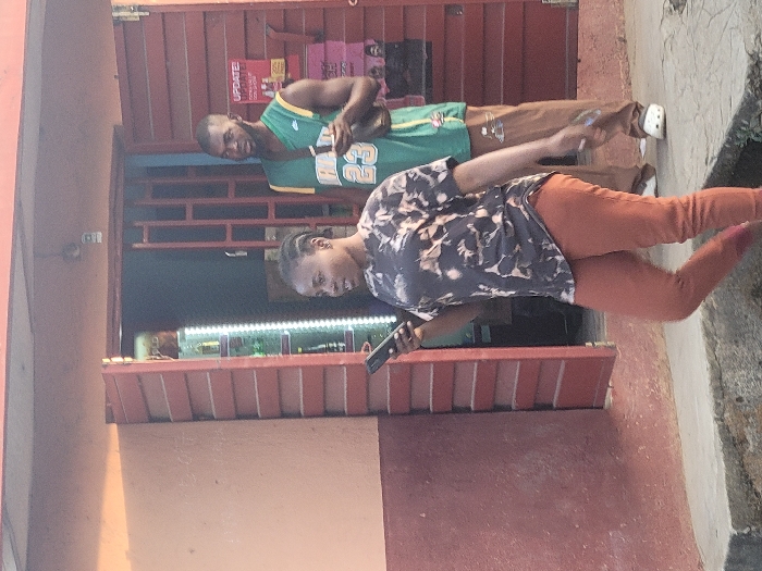 Two individuals stand outside a store. A man wearing a green basketball jersey and tan pants is behind a woman who holds a black object (likely a phone or device) in her hand. She sports a patterned dark top and orange pants. The store has a red facade with a barred window display and promotional posters on top. The ground appears to be concrete, and the overall setting seems to be a casual street scene.