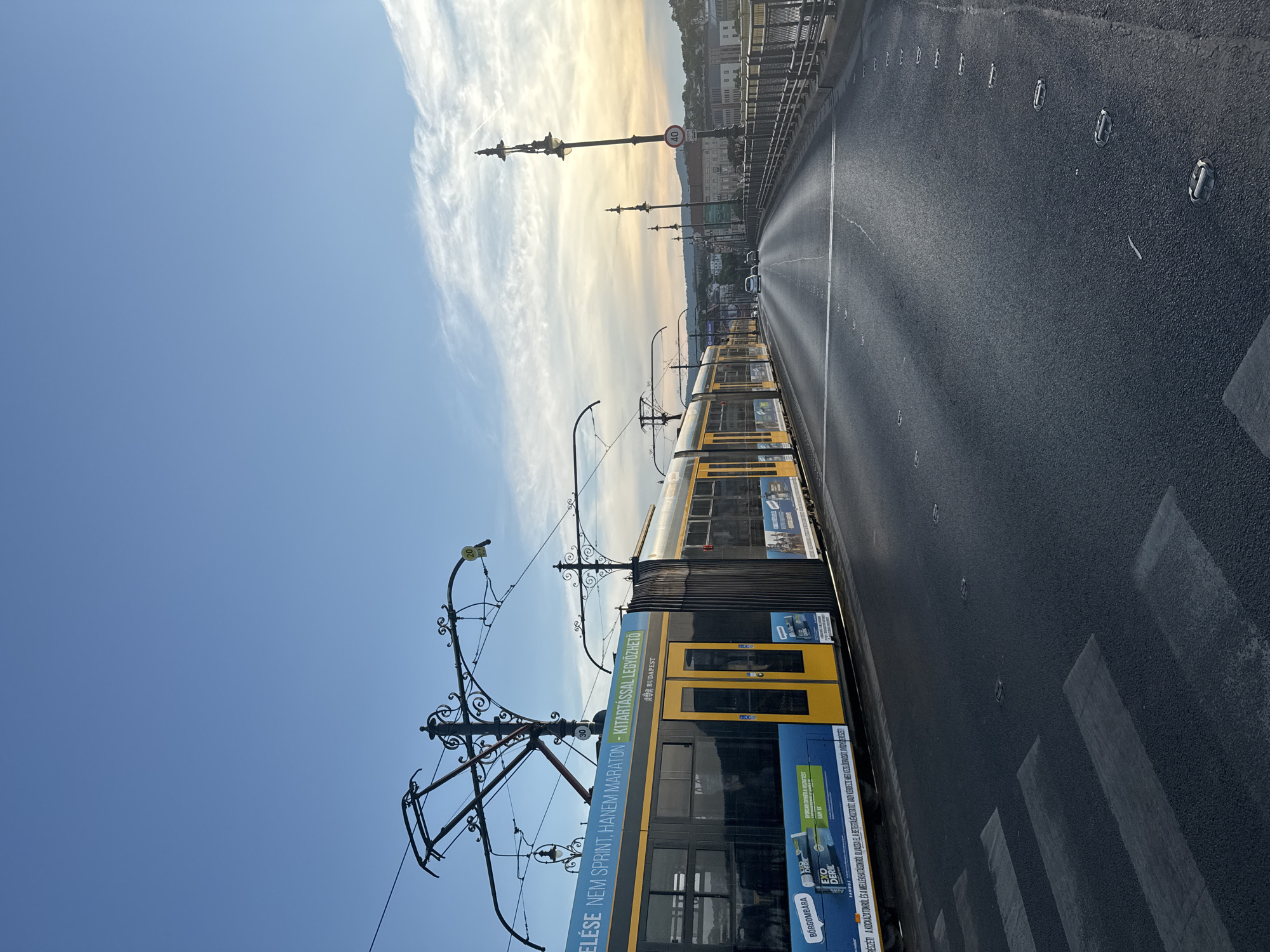 This image depicts a bright, sunny day in Budapest, Hungary, likely during the late afternoon or early evening as suggested by the warm light and long shadows. The perspective is from a slightly elevated viewpoint, looking down a road that appears to be a bridge.
The most prominent object in the foreground is a yellow and blue tram, its side adorned with advertisements. The tram's overhead electrical wires and supporting structure are also clearly visible, stretching across the frame. To the right of the tram is a wet asphalt road with a pedestrian crossing marked by white stripes. Further down the road, several cars are visible, suggesting moderate traffic.
In the background, a line of buildings and structures stretches across the horizon, hinting at the cityscape of Budapest. Ornate lampposts line the side of the road, some with "40" and "30" speed limit signs attached. The sky is a vibrant blue, with wispy white clouds catching the golden light of the setting sun.
The overall scene conveys a sense of urban movement and daily life, bathed in the picturesque light of the golden hour. The visible text on the tram includes "BUDAPEST" and promotional slogans in Hungarian, such as "ELÉSE NEM SPRINT, HANEM MARATON - KITARTÁSSAL LEGYŐZHETŐ" which translates to "Not a sprint, but a marathon - conquerable with endurance". Other text appears to be advertising product names like "EXO DERIL".