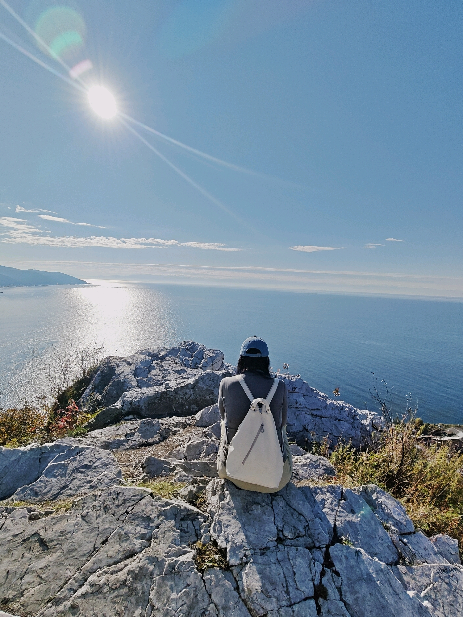 A lone individual is seated on a rocky outcrop overlooking a vast expanse of ocean under a clear blue sky. The sun, positioned high and to the left of the frame, casts a brilliant glare on the water and creates lens flare effects in the sky. The person, viewed from behind, wears a dark top, a blue baseball cap, and a light-colored backpack. The rocky terrain is rough and textured, with patches of dry grass and some sparse vegetation. In the distance, a hazy coastline with a faint town or settlement is visible. The overall impression is one of peaceful contemplation and appreciation of a scenic natural landscape. There is no information in the image to confirm or deny the location being Baikal, Russia.