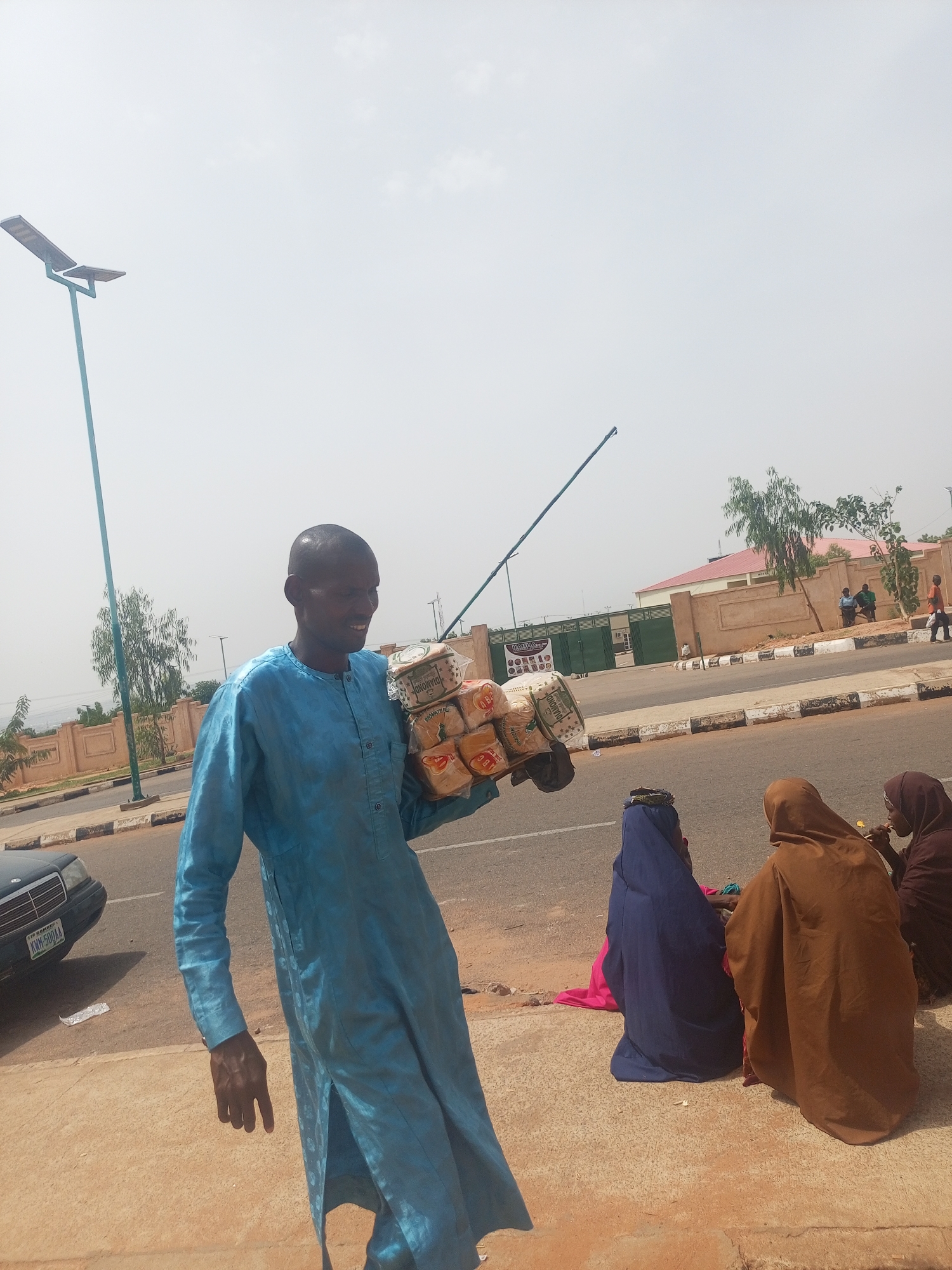 The image depicts a vibrant street scene in Gombe, Nigeria, on a bright, sunny day. The main subject is a man walking in the foreground, carrying a large stack of individually packaged loaves of bread on his left arm. He is dressed in a light blue, long-sleeved traditional garment and has a slight smile on his face, looking towards the right side of the frame. The bread packages display text such as "URONOYOL" and "DIAMOND."
To the right of the man, seated on the reddish-brown ground beside the road, are three women. Two are more prominent, dressed in flowing traditional attire, one in a brown hijab/abaya and the other in a dark blue one, with their backs partially turned to the viewer. A third woman in a darker garment is partially visible next to them.
The scene features a paved road with a distinctive white and black striped curb. On the left side of the road, a black car, possibly a Mercedes-Benz, is parked, displaying a Nigerian license plate "KWM-500AA" and "GOMBE STATE" clearly visible. A tall, blue-green pole with a solar panel and a street light head extends into the sky from the left side of the frame. Another similar pole is visible further in the background on the right.
In the mid-ground and background, there are buildings with light-colored walls and green fences, suggesting residential or commercial properties. Sparse green trees and shrubs are scattered throughout the landscape, with hills faintly visible in the far distance under a clear, bright sky. The overall activity suggests everyday life, with the man acting as a street vendor and others engaging in daily routines.