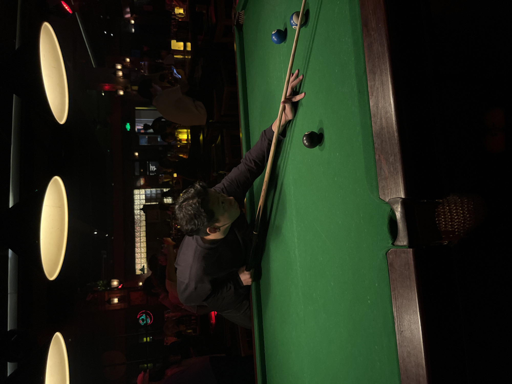 A medium shot depicts a person playing pool indoors. The individual is positioned on the left side of the frame, leaning over a pool table covered in green felt. They are holding a pool cue with their right hand extended towards the center of the table, and their left hand providing support. Two pool balls, one blue with white spots and the other black, are visible on the table's surface.
The background is dimly lit, suggesting a bar or entertainment venue. Vertical, elongated light fixtures are mounted on a wall to the left. Other people can be seen in the background, but they are out of focus.
The environment suggests an indoor recreational space. The image captures a moment of concentration and action during a game of pool. There is no specific indication that the location is in Liangjiang, China; therefore, it cannot be determined with certainty.