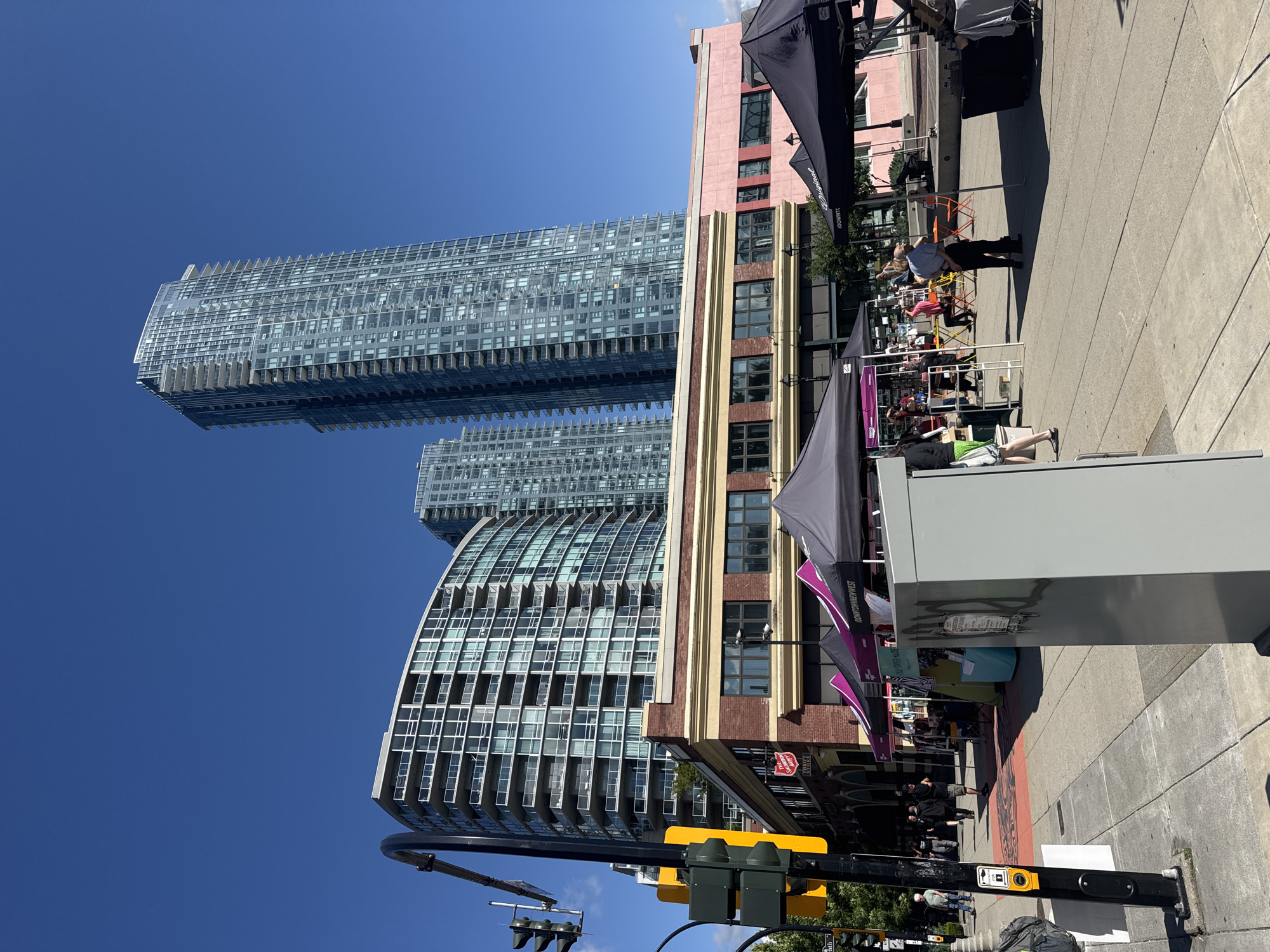 In New Westminster, Canada, a street scene unfolds under a clear blue sky. In the background, several modern, glass-fronted high-rise buildings pierce the sky, one of them being a particularly tall, slender skyscraper. In the midground, a low-rise brick building with arched windows features a prominent Salvation Army Thrift Store sign. In front of this building, a row of market tents, some purple and some black, are set up, suggesting an outdoor market. People are visible browsing the stalls and walking along the sidewalk. To the left, a traffic light pole with a pedestrian crossing signal stands at the intersection. The foreground is dominated by a grey utility box and a paved sidewalk. The overall impression is a sunny day in a city with a mix of historic and contemporary architecture, bustling with activity at a street market.