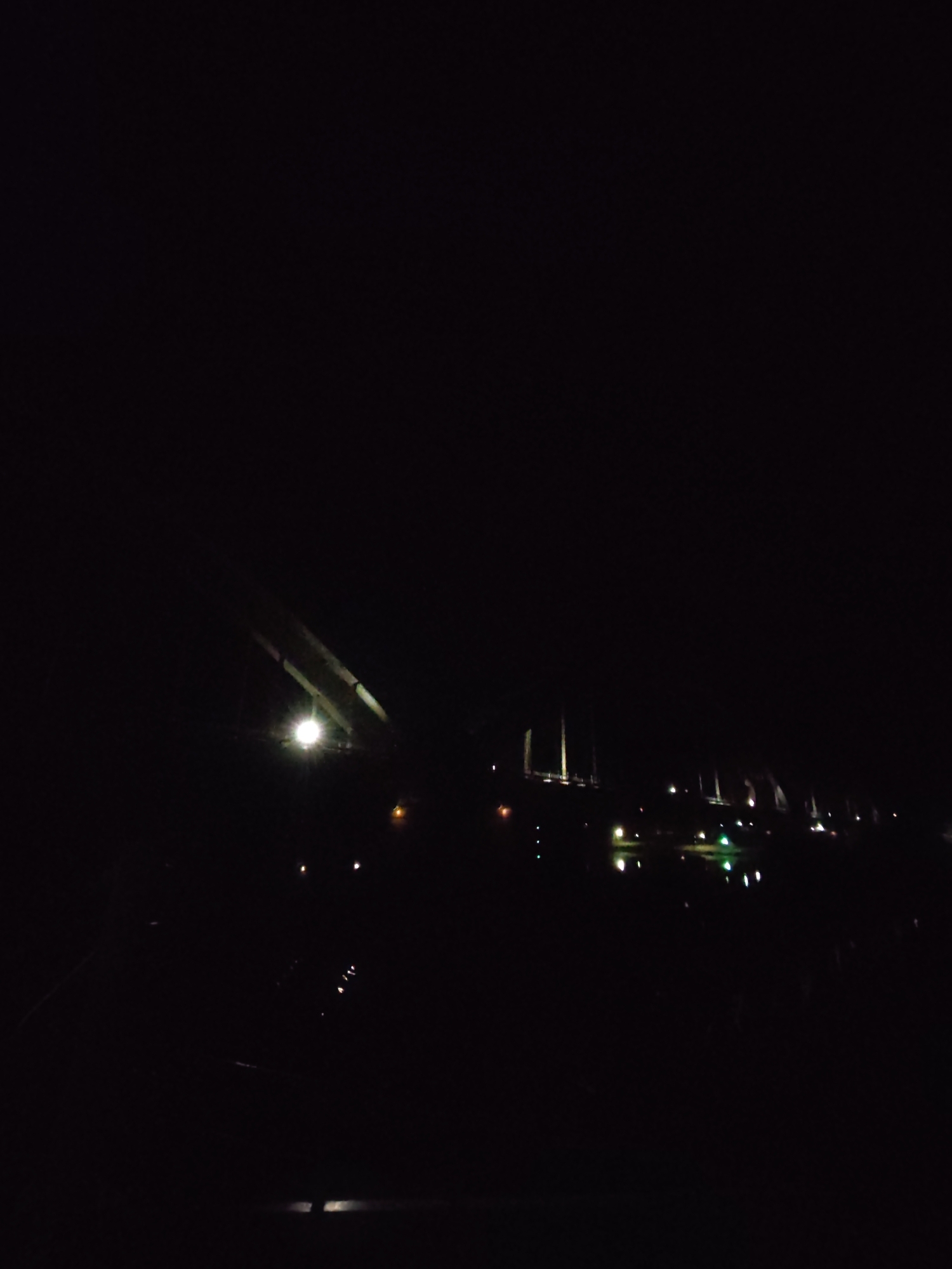 The image appears to be taken at night in Gladyshevo, Russia. A bridge is visible in the middle distance, illuminated by several lights. Some of these lights are reflected in a body of water. The foreground is dark, obscuring any details. The overall scene is poorly lit, with most of the image consumed by darkness.