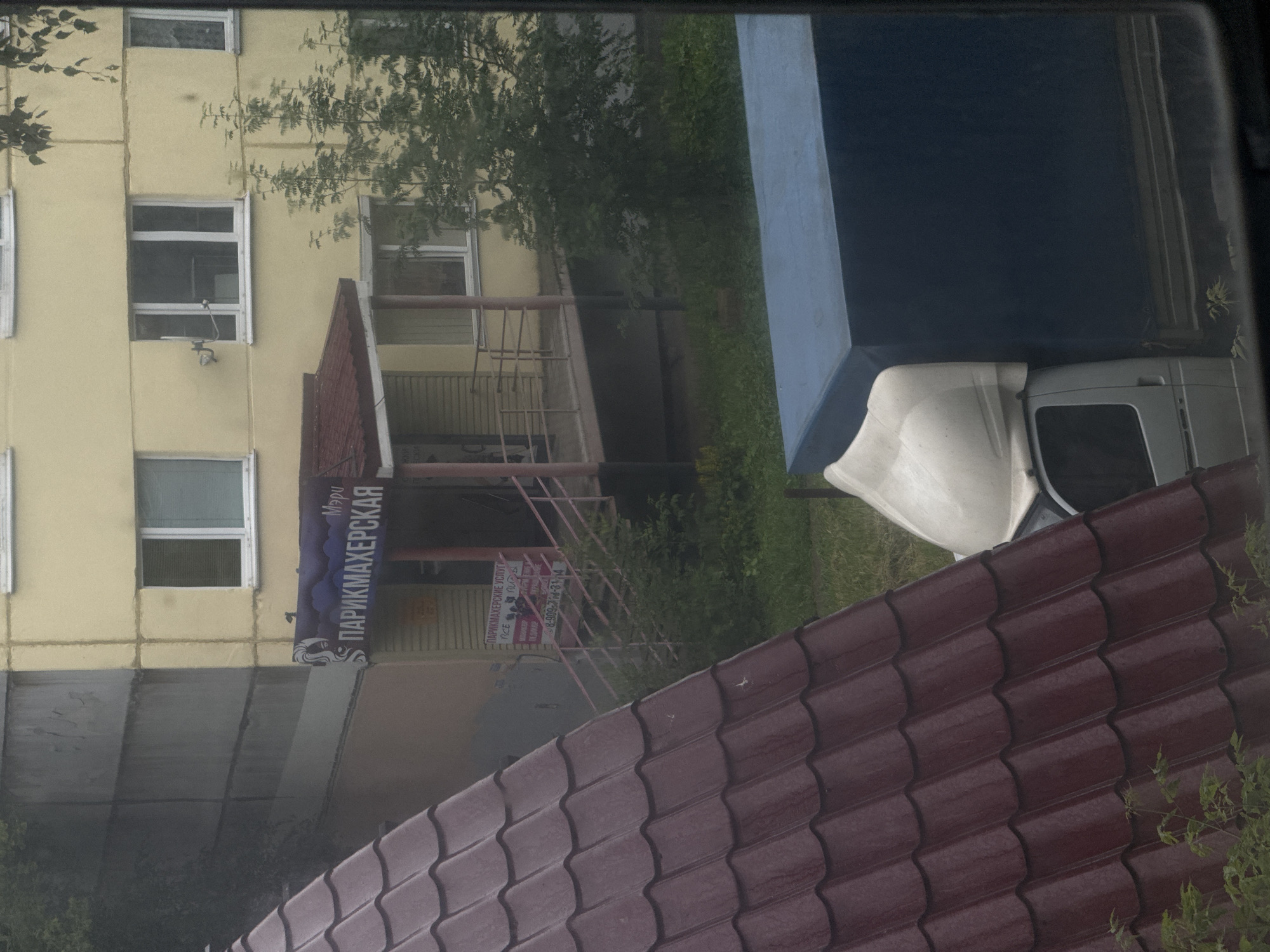 The image captures a view of a street or courtyard in Izhevsk, Russia, dominated by urban architectural elements and a parked truck. In the foreground, a dark red, corrugated metal roof slopes diagonally across the frame, suggesting a building or structure with a sloped roof. To the left, a lighter gray, weathered concrete wall of a building is visible.
Behind these foreground elements, a light yellow apartment building with multiple windows is the central architectural feature. Some windows have blinds or curtains, and one window on the upper floor has a visible satellite dish mounted to its exterior. A small, dark blue sign with white lettering reading "Мэру ПАРИКМАХЕРСКАЯ" (Mayor Hairdresser) is affixed to the lower part of the yellow building, indicating the presence of a business. Below this sign, another, smaller pink sign with Russian text and a phone number is partially visible, likely advertising services.
The scene is set against a backdrop of green foliage, including trees and bushes, suggesting a somewhat vegetated urban environment. In the lower right portion of the image, the rear section of a white truck with a large blue tarp-covered cargo area is parked. The truck's cab is white, and its windshield is visible. The overall impression is a mundane, everyday scene in a residential or commercial area of Izhevsk.