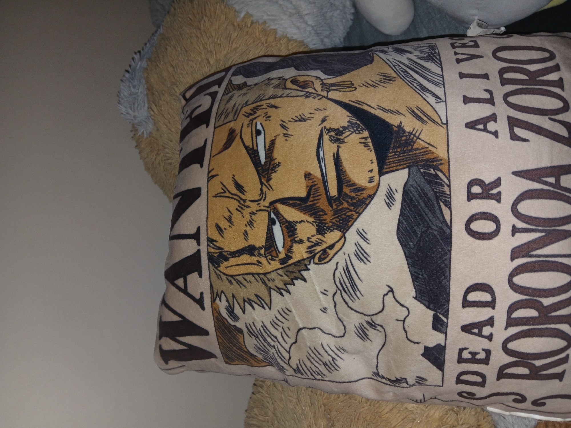 The image shows a plush pillow resting on a tan stuffed animal. The pillow depicts the wanted poster of Roronoa Zoro, a character from the Japanese manga series One Piece. The poster image includes his face and the text "WANTED", "DEAD OR ALIVE", and "RORONOA ZORO". No information related to Bengaluru, India is visible.