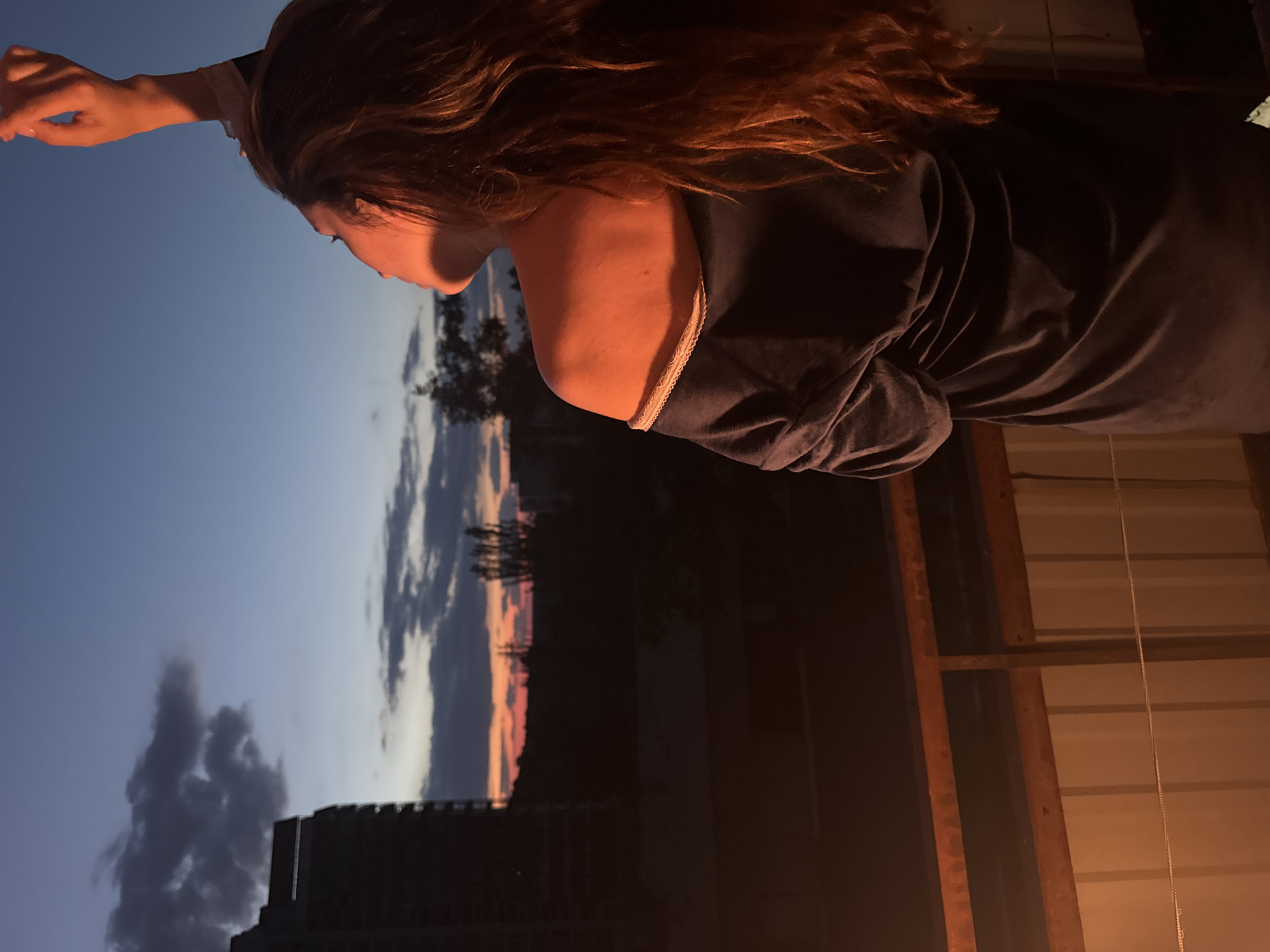 A woman stands on a balcony facing away from the camera, looking up at a twilight sky. Her right arm is raised, and her hand is extended upwards. The sky is a gradient of deep blue at the top, transitioning to shades of orange and pink near the horizon, with scattered clouds. In the background, silhouetted against the sky, are buildings, trees, and the vague outline of a city. The lighting on the woman suggests the warm glow of the setting sun or artificial light. The balcony railing and a corrugated metal wall are visible in the foreground, suggesting an urban setting, possibly in Odesa, Ukraine. The woman has long, reddish-brown hair that cascades down her back. She is wearing a dark, off-the-shoulder garment that appears to be made of a soft material. The overall impression is one of contemplation or reaching for something.