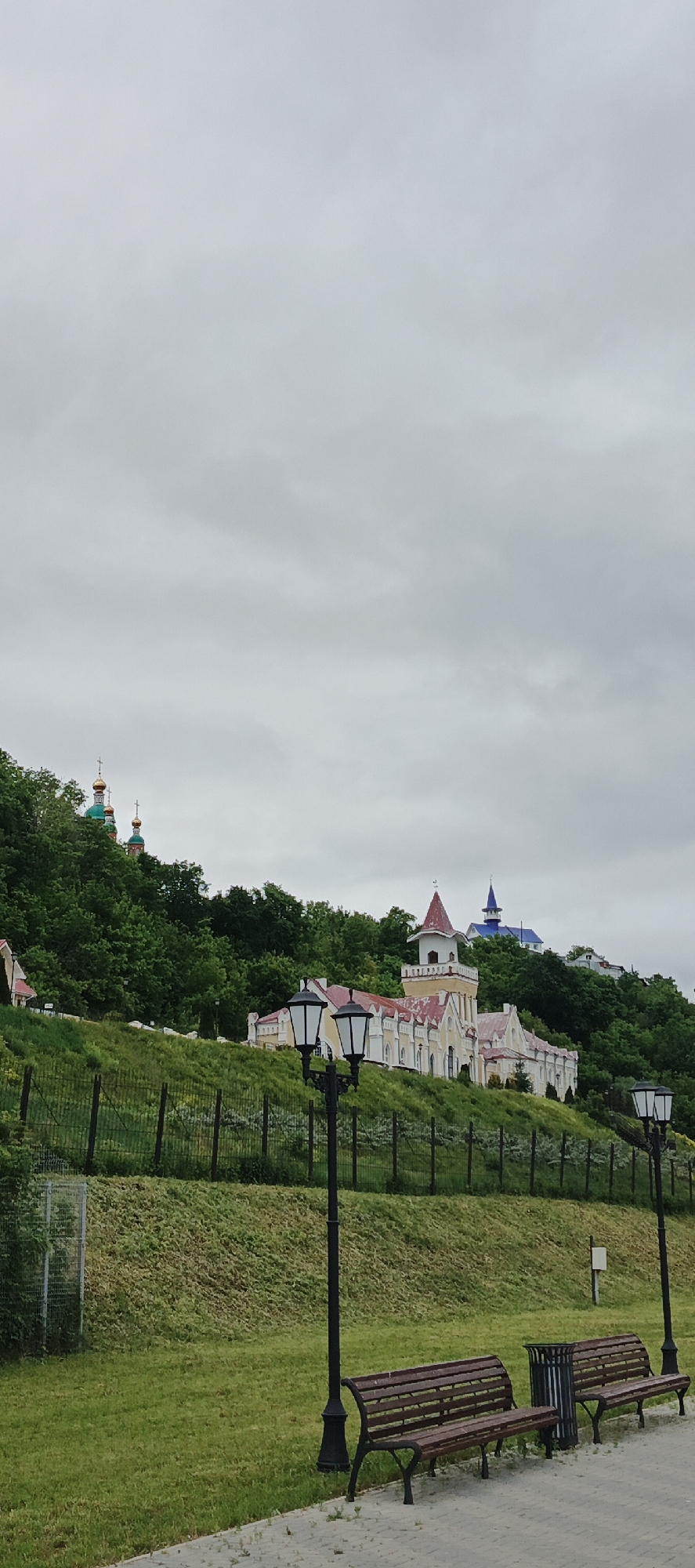 This full-shot vertical image depicts a panoramic view of a park-like setting in the city of Sarapul, Russia, under an overcast sky. In the foreground, two dark brown slatted park benches with black metal frames are positioned on a light grey paved walkway, running horizontally across the bottom of the frame. To the left of the benches, a dark grey cylindrical trash receptacle is visible. Two ornate black lampposts with multiple white glass lanterns stand upright, one to the left of the benches and one to the right, illuminating the paved area. Adjacent to the walkway is a strip of neatly maintained green grass.
Behind the foreground elements, a steep, grassy hill rises, covered with dense green trees towards its summit. A dark mesh fence runs along the base of the hill, separating the lower park area from the slope. Mid-slope on the right side, a prominent, light yellow building with multiple gabled sections and a striking red roof is visible. This building features a distinctive central tower with a pointed red roof. Further up the hill and to the left, partially obscured by trees, two traditional Orthodox church domes, green with gold accents and crosses, are visible. To the right, near the crest of the hill, another building with a pointed blue roof or spire can be seen peeking above the tree line. The upper half of the image is dominated by a uniform, light grey, cloudy sky. No people are present in the scene.