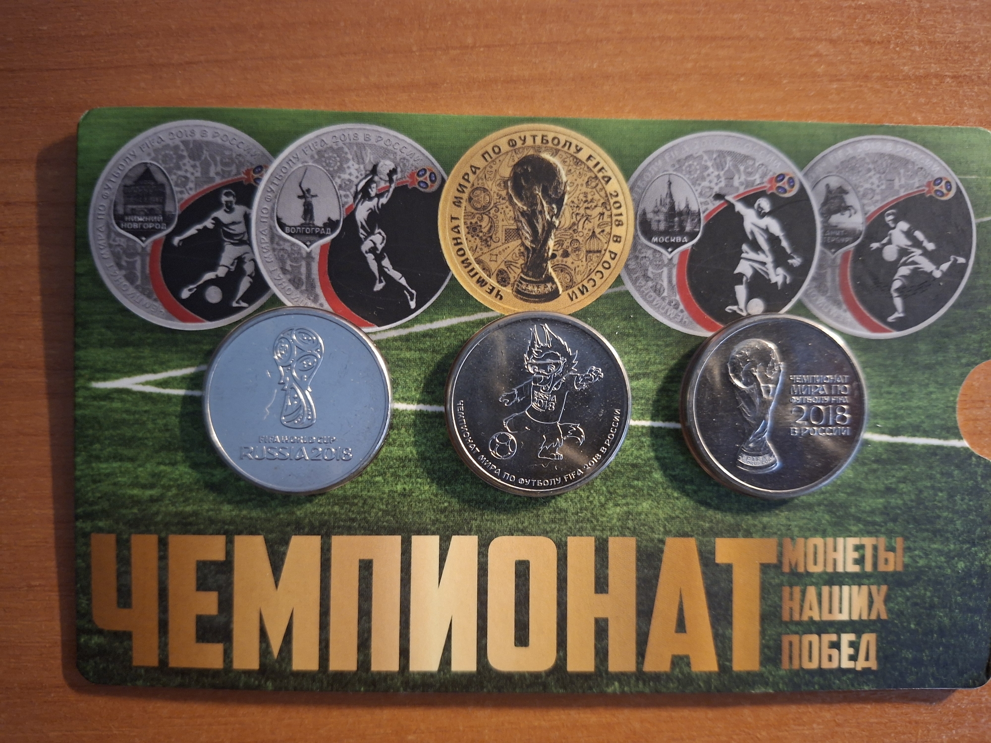 The image displays a collection of coins laid out on a green surface designed to resemble a soccer field. The coins are positioned on the right side of the image, while a large vertical text is present on the left. The text is in large, bold, orange letters and reads "ЧЕМПИОНАТ НАШИХ МОНЕПЫ ПОБЕДА". The coins themselves appear to be commemorative medals related to the FIFA World Cup 2018 in Russia. Several coins are visible, some featuring images of soccer players in action, while others display iconic landmarks. One prominent gold-colored coin prominently features a stylized representation of the FIFA World Cup trophy. The background is a wooden surface, indicating the items are placed on a table or similar surface. There are no people visible in the image, and the focus is solely on the coins and the printed material.