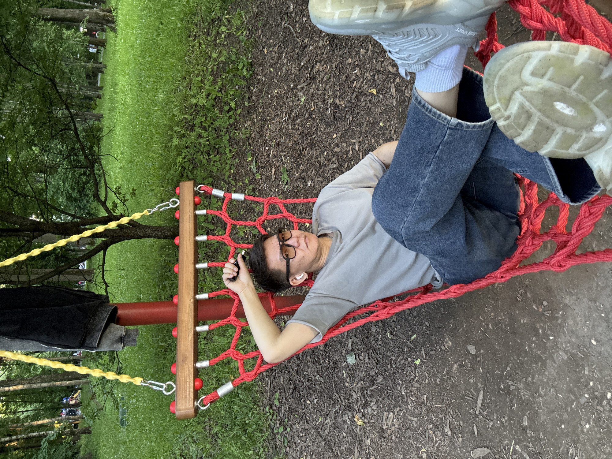 The image, rotated to show the scene correctly, depicts an individual relaxing in a red rope hammock swing within a park in Moscow, Russia. The person, an adult, is lying on their back, looking towards the viewer with a neutral, relaxed expression. They are dressed in a light gray short-sleeved t-shirt, dark blue jeans, white socks, and light gray sneakers. White wireless earbuds are visible in their ears, and they wear dark-rimmed glasses. A small black object, possibly a phone or vape pen, is held in their right hand.
The red rope hammock, intricately woven, is suspended from a sturdy red pole. This pole also features several wooden crossbars adorned with small red spherical elements. A yellow rope or chain is attached to the pole, and a dark-colored jacket is draped over it. The ground immediately beneath and around the hammock is covered with dark wood chips or mulch. Beyond this, the background reveals a vibrant green grassy area lined with numerous tall trees, indicative of a natural park environment. The lighting is soft and even, suggesting either an overcast sky or indirect sunlight during daytime hours. On the sole of one of the sneakers, the text "KGA SPORT" is visible. The overall scene conveys a sense of leisure and peaceful recreation.