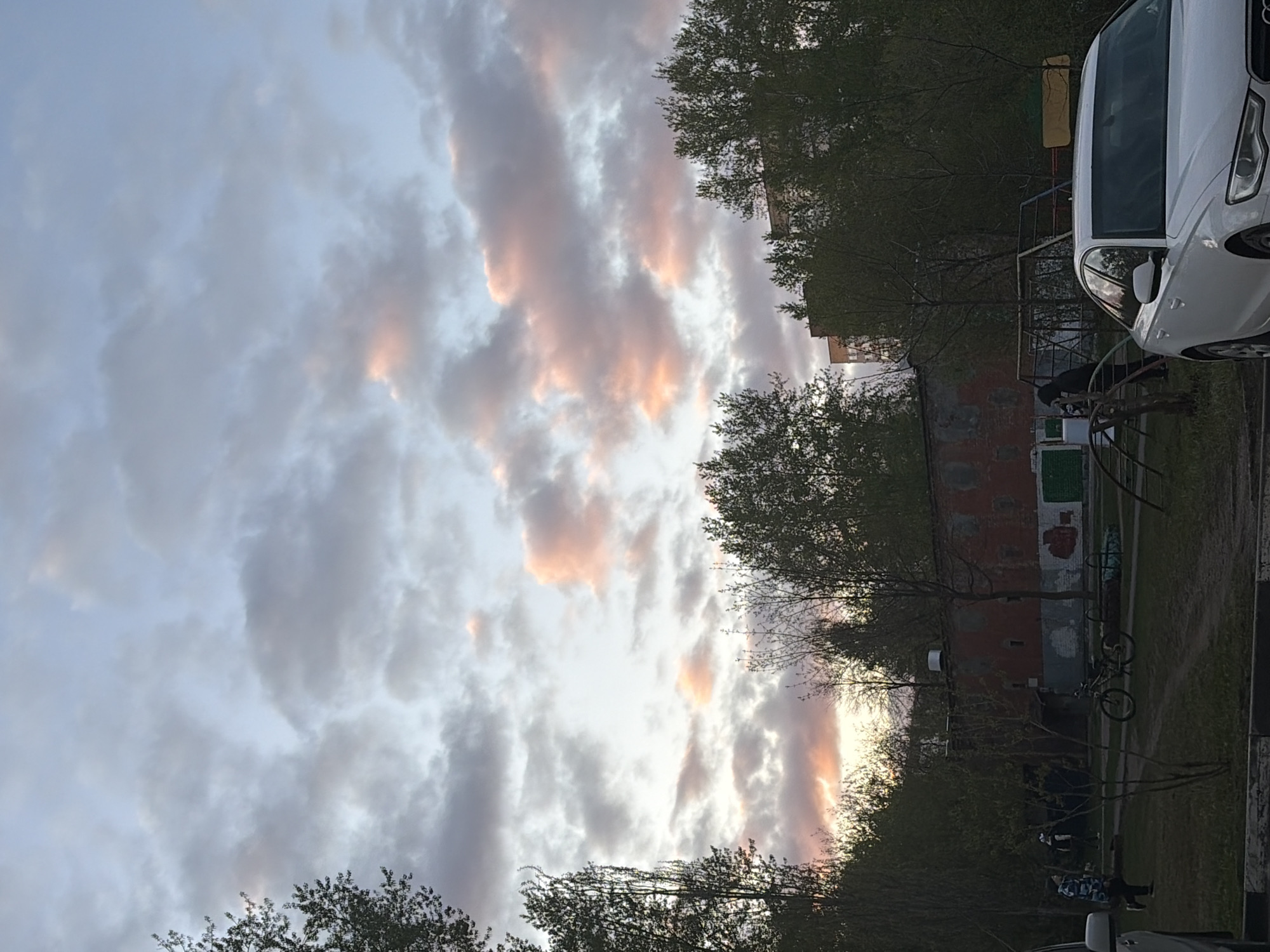 The image depicts an outdoor scene in Perm, Russia, at dusk. The sky is filled with predominantly gray clouds, some tinged with shades of orange and pink near the horizon. Trees with budding leaves are visible in the foreground and midground, lining the edges of what appears to be a residential area. A white car is parked on the right side of the frame. A brick building with visible graffiti is situated in the midground, next to a playground structure and a bicycle. Several people are present near the building and playground, some possibly seated. The overall environment suggests a typical urban neighborhood.