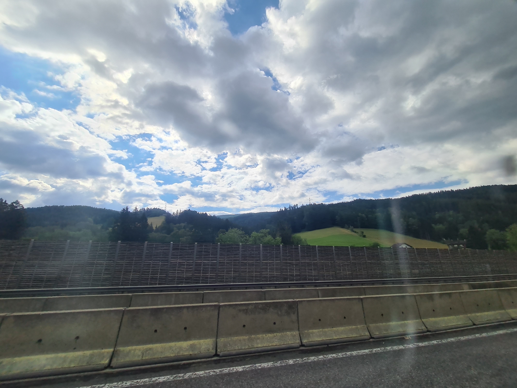 The image depicts a view from a highway in Spital am Semmering, Austria. In the foreground, there are concrete barriers separating the lanes of the highway. Behind the barriers, there is a dark wooden sound barrier. Beyond the sound barrier, rolling green hills are visible, dotted with trees. The sky is partly cloudy with patches of blue sky interspersed with large, dramatic clouds. In the distance, more hills covered in dark green trees form the backdrop, with a few buildings or houses visible nestled among them. The overall scene suggests a scenic drive through a mountainous or hilly region of Austria.