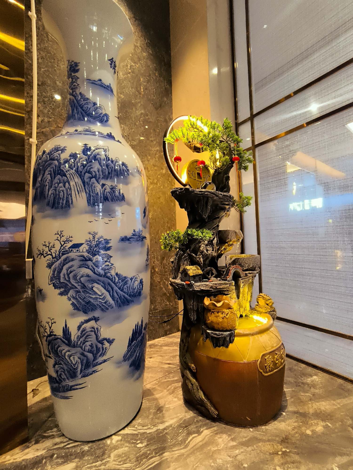 The image depicts an interior scene, likely a room or lobby, with an emphasis on decorative elements. On the left, a large, ornate porcelain vase with blue and white landscape patterns dominates the foreground. To its right stands a miniature water fountain adorned with artificial greenery, small red lanterns, and a golden pot at its base, which is illuminated. A golden toad figure sits atop the pot. The fountain appears to be a traditional Chinese-style piece. The background features a textured wall and a window covered by a light-colored, translucent blind. The flooring is a marbled surface. There are no people visible in the image. The scene is well-lit, highlighting the decorative objects. The image does not contain any information that would allow for identification of its specific location within the city of Ürümqi, China.