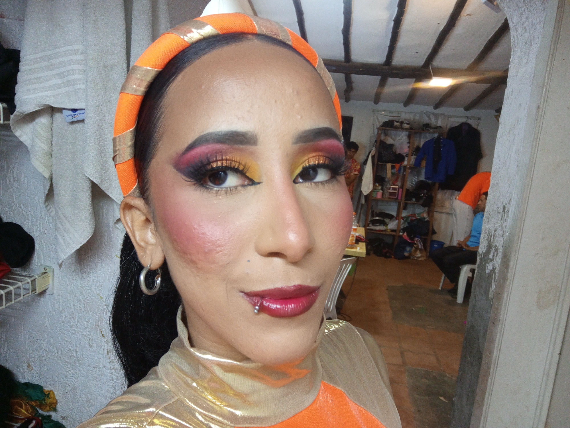 A close-up shot depicts a woman with dark hair pulled back, wearing a bright orange headband with gold accents, noticeable makeup including red-pink blush, heavy eyeshadow, and deep red lipstick with a lip piercing on the lower left side. She is wearing a metallic gold and orange top. The background reveals an interior space with a tiled floor and a partially visible individual seated on a white chair, wearing orange and blue clothing. A clothing rack with hanging garments is also visible in the background. The location appears to be an indoor area, possibly a dressing room. The exact location in Porlamar, Venezuela is not discernible from the image.