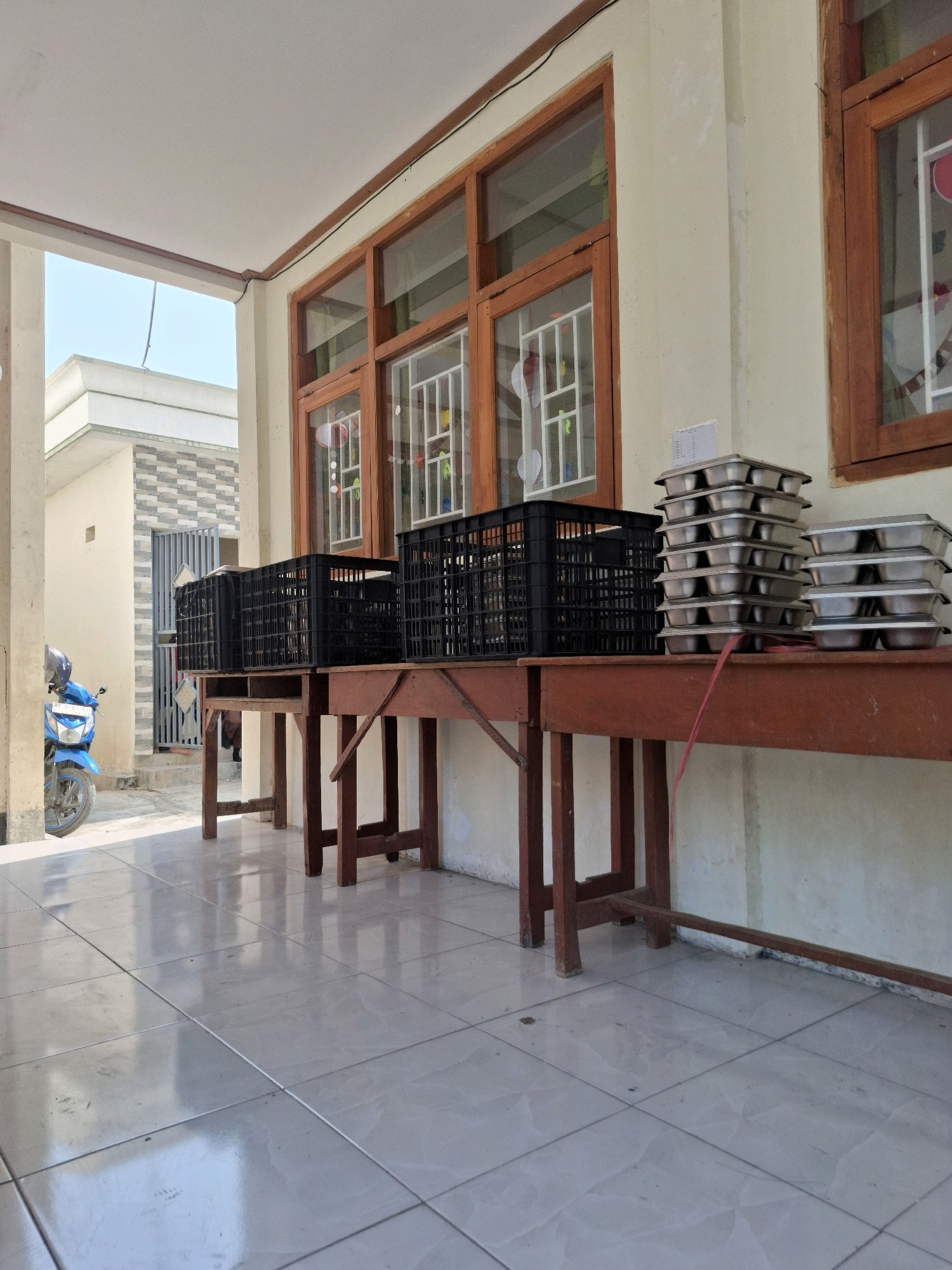 The image depicts an outdoor scene in front of a building, likely located in Pilangkenceng, Indonesia. The foreground is dominated by a tiled floor reflecting the bright sunlight. Two wooden tables are positioned on the tiles, one slightly further back and appearing more substantial.
On the tables, there are several black plastic crates and stacks of metal trays. The crates appear to be empty. The metal trays are stacked neatly, with some possibly containing items, though their contents are not clearly discernible. A red string or ribbon is visible draped across the front table, near the stacked trays.
Behind the tables, the building features large wooden-framed windows with decorative white grilles. The walls of the building are light-colored, possibly cream or beige. To the left of the tables and building, a blue motorcycle is parked, with its front wheel visible and a helmet resting on the handlebars. Further in the background, a gate and part of another building are visible, suggesting a residential or institutional setting. The overall impression is one of an area used for storage or perhaps a temporary setup for an event. The lighting indicates it is daytime, with strong shadows cast by the tables and building.