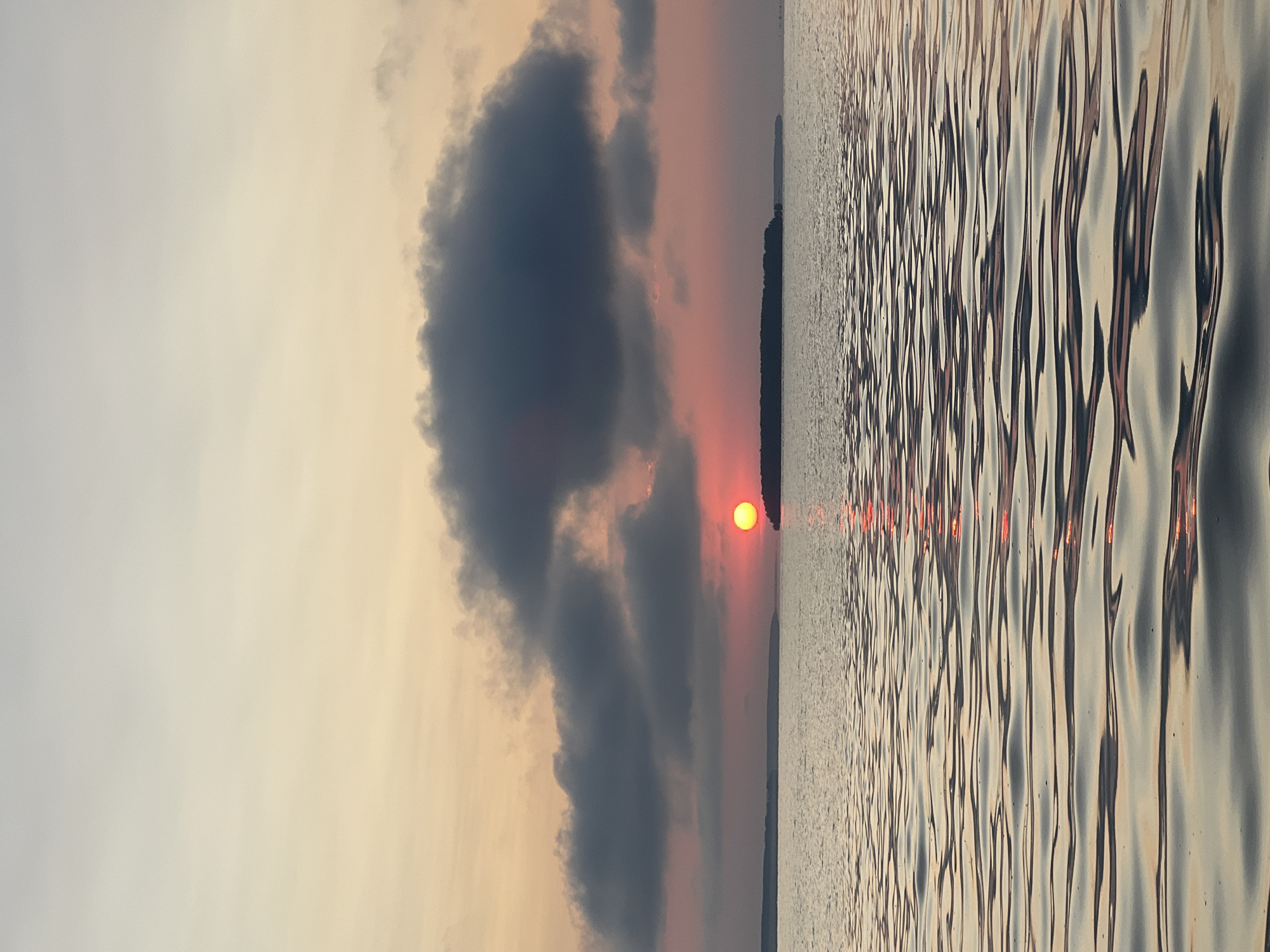 A serene sunset over a body of water near Novopichugovo, Russia. The sun, a vibrant orange orb, is partially obscured by dark clouds, casting a warm glow across the sky. The water's surface is rippled, reflecting the colors of the sunset. A narrow strip of land, possibly an island or coastline, is visible in the distance. The scene evokes a sense of tranquility and natural beauty.