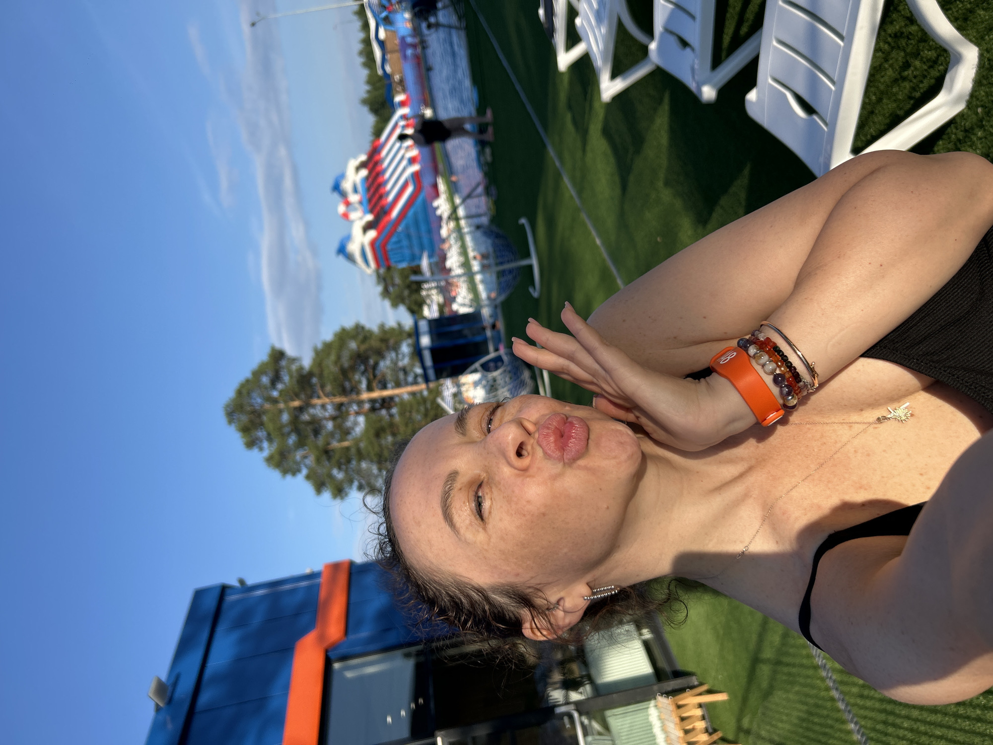 The media file shows a woman posing for a selfie in an outdoor setting. She is leaning back, with her face angled towards the camera, making a kissing gesture. Her hair is dark and curly, and she has freckles across her face. She is wearing a black top and several bracelets on her left wrist, one of which is bright orange. A delicate necklace with a star pendant rests on her collarbone. Her right hand is raised to her face, touching her cheek.
In the background, a large inflatable water slide with red, white, and blue sections is visible, suggesting a recreational area. White lounge chairs are set up on a green, artificial turf surface. To the left, a portion of a blue and orange building is visible. Tall pine trees are present in the distance, and the sky is a clear, bright blue, indicating it is daytime with fair weather. The overall impression is one of leisure and fun on a sunny day. The context provided states the location is Votkinsk, Russia. No visible text is present in the media file.