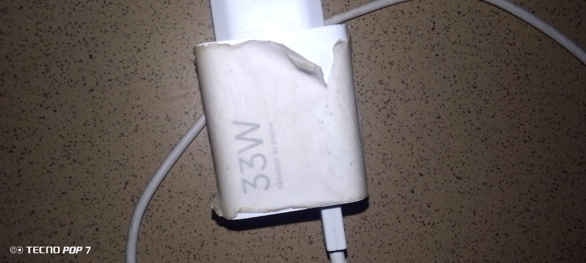 An overhead, close-up shot shows a white 33W Xiaomi charger plugged into a white cable. The charger's white sticker is peeling at the top and is soiled. The charger and cable are on a speckled, brown surface. The bottom left corner of the image displays the text "TECNO POP 7".