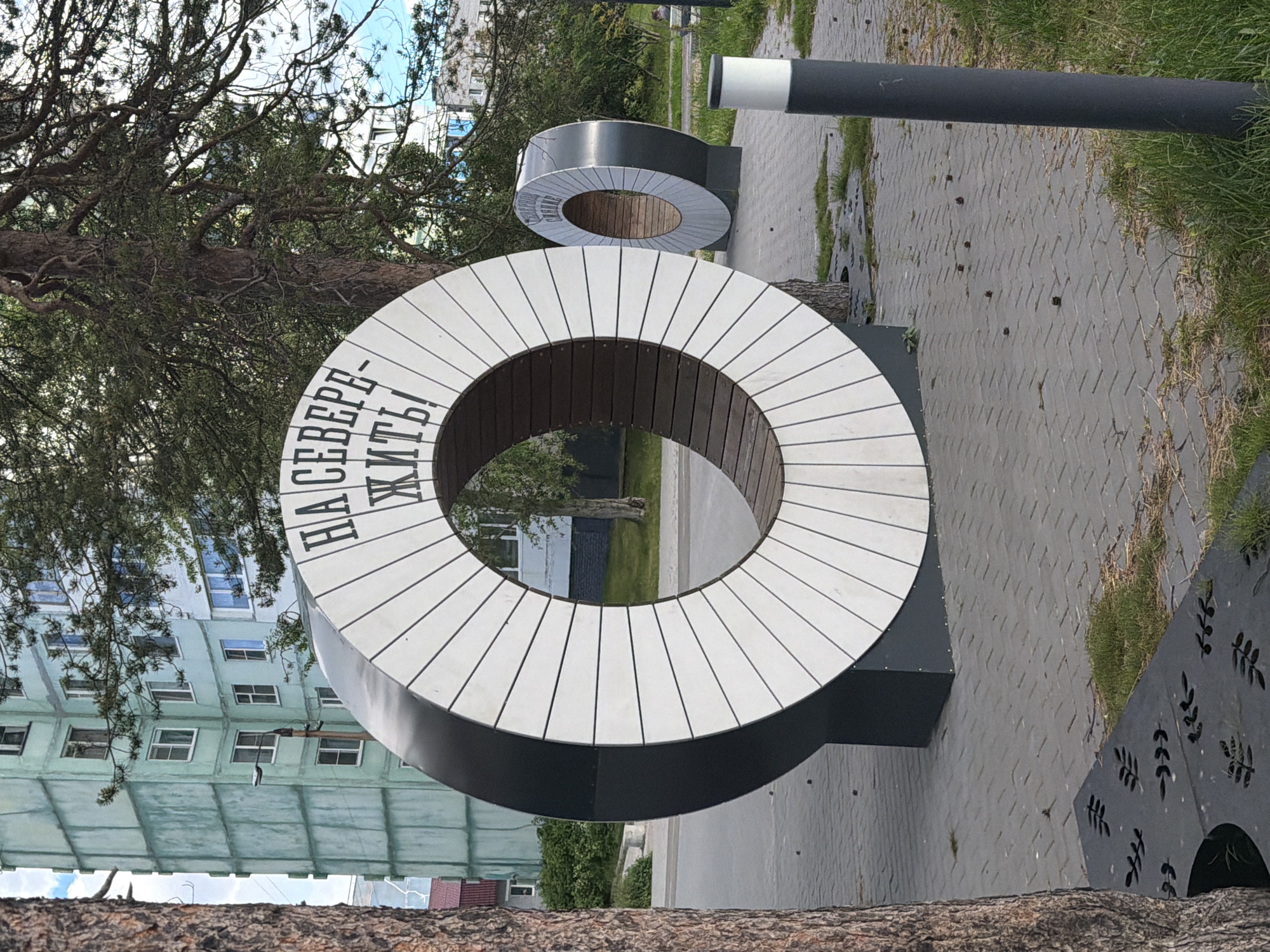 A circular bench with the Russian text "НА СЕВЕРЕ ЖИТЬ!" (To Live in the North!) is the main subject in this outdoor scene. The bench is white on the outside with wooden planks on the inside and is situated on a paved area. Behind the bench, a light blue apartment building stands, suggesting an urban or suburban setting in Russia, specifically Polarnie Zori. The scene is framed by the branches of a large tree in the foreground. The lighting suggests it is daytime. There is another smaller, similar circular bench visible in the background. A dark grey pole with a white stripe runs horizontally across the upper right portion of the image. The ground is covered with grey paving stones and patches of grass. Decorative metal panels with leaf cutouts are also visible on the right side of the image.
