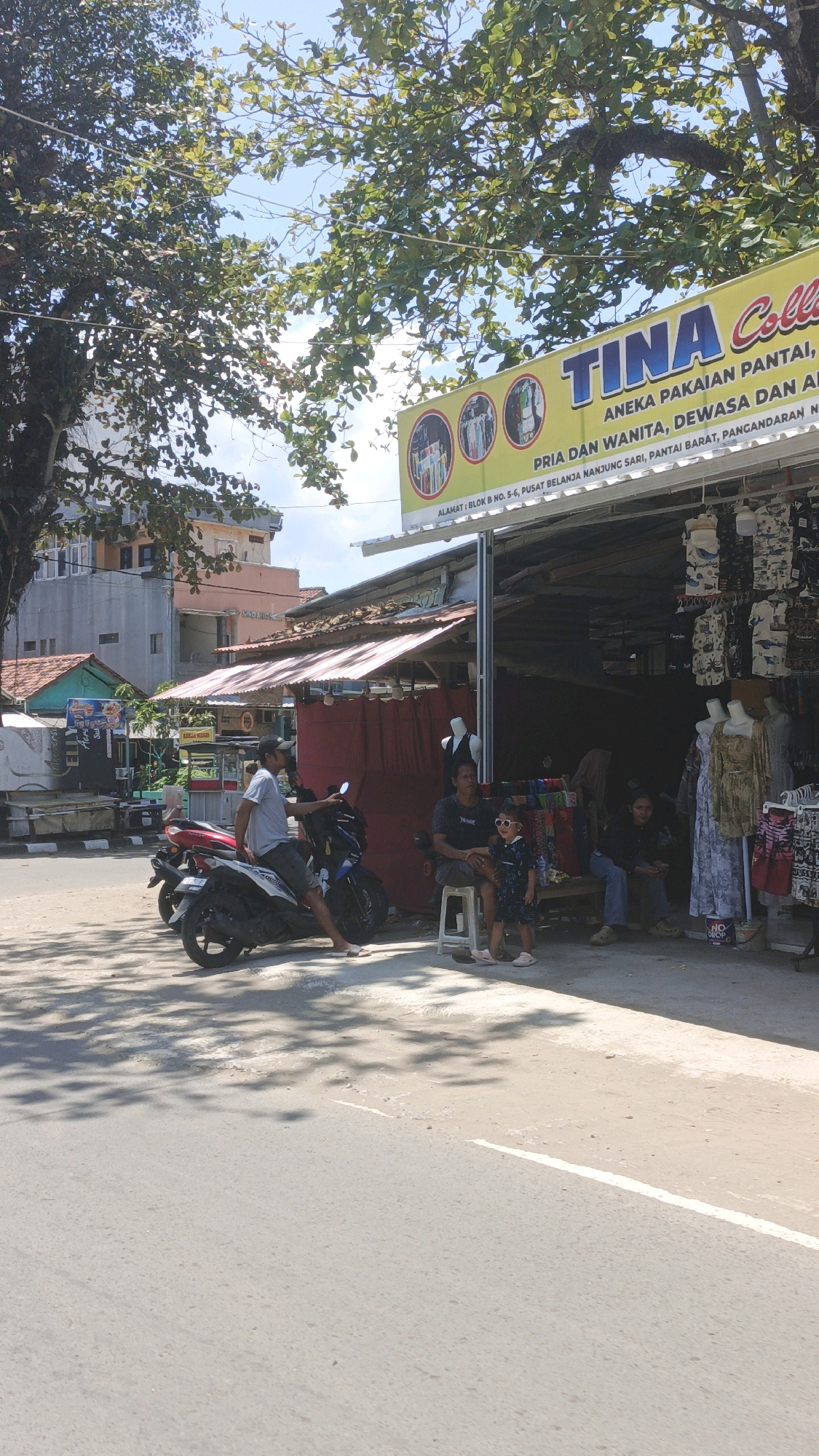 The image captures a street-side scene in Desa Pagandaran, Indonesia, dominated by a shop with a bright yellow sign that reads "TINA Collec" and "ANEKA PAKAIAN PANTAI, PRIA DAN WANITA, DEWASA DAN AN..." and the address "ALAMAT : BLOK B NO. 5-6, PUSAT BELANJA NANJUNG SARI, PANTAI BARAT, PANGANDARAN N". The shop appears to sell beachwear, as indicated by the signage and the racks of clothing visible outside.
In the foreground, two motorcycles are parked on the paved road. A man wearing a grey t-shirt and shorts is seated on one of the motorcycles, facing towards the shop. To the right of the motorcycles, three people are seated near the shop entrance. An adult woman with dark hair is sitting on a white stool. Next to her, a young girl wearing sunglasses and a dark patterned dress is standing and looking towards the shop. Further to the right, another adult is seated on a dark stool.
The shop itself is open-fronted with a corrugated metal roof supported by posts. Racks of colorful clothing, primarily t-shirts with nautical or beach-themed prints, are displayed outside the shop. A mannequin wearing a dark outfit is visible inside the shop, and other clothing items and fabrics are stacked behind it. To the left of the shop, another stall with a red awning is visible, displaying more merchandise.
In the background, buildings of varying architectural styles and colors line the street, including a multi-story building with a pale facade and orange accents. Lush green trees with broad leaves provide shade over the scene, with their branches extending towards the bright blue sky. The ground is a mix of paved road and a lighter-colored walkway or dirt area in front of the shops, where shadows from the trees are cast.