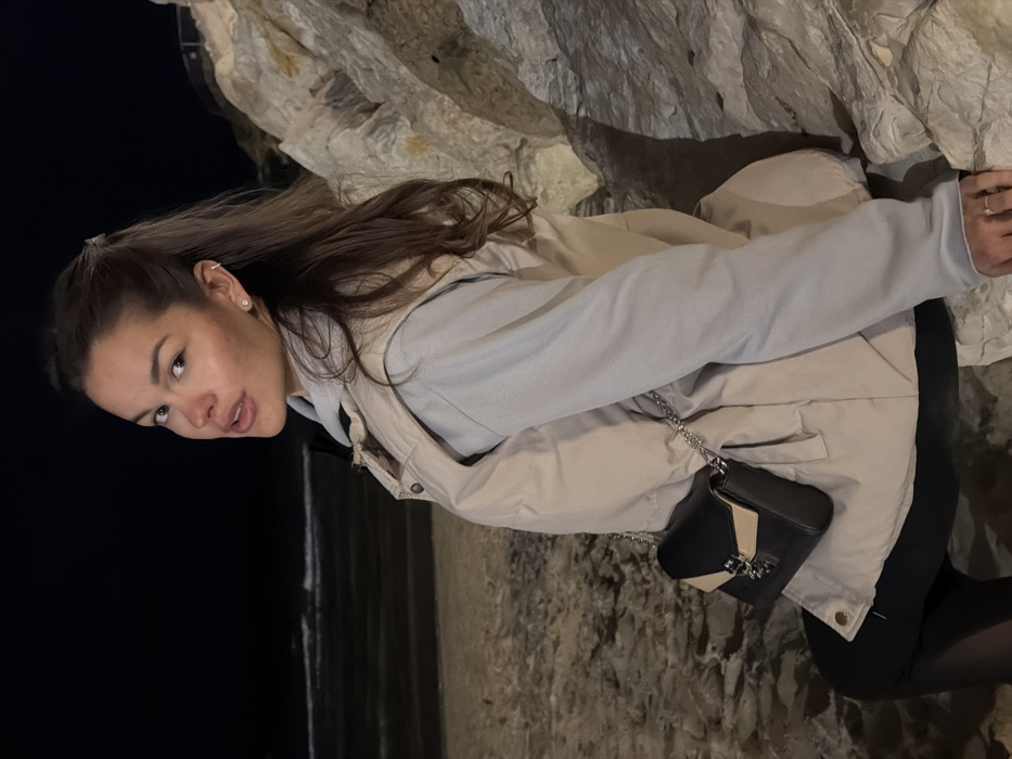 The image captures a female individual at night on a Tel-Aviv beach, specifically positioned among a formation of light-colored rocks along the coastline. The subject, a young adult with long, dark brown hair styled in a half-ponytail, is seated or crouched against these rocks on the right side of the frame, facing the camera with a slight turn over her right shoulder. She is dressed in a light grey long-sleeved top, overlaid with a beige puffer vest, and dark leggings. A black and beige two-tone shoulder bag with a silver chain strap is worn across her body. Noteworthy details include a stud earring in her left earlobe, a helix piercing in her left ear, and a ring on her right ring finger. To the left, a sandy beach transitions into the dark waters of the Mediterranean Sea, where subtle waves are visible against the night sky. The scene is illuminated by artificial light, highlighting the subject and the immediate rock formations, while the background and sea remain largely in shadow.
