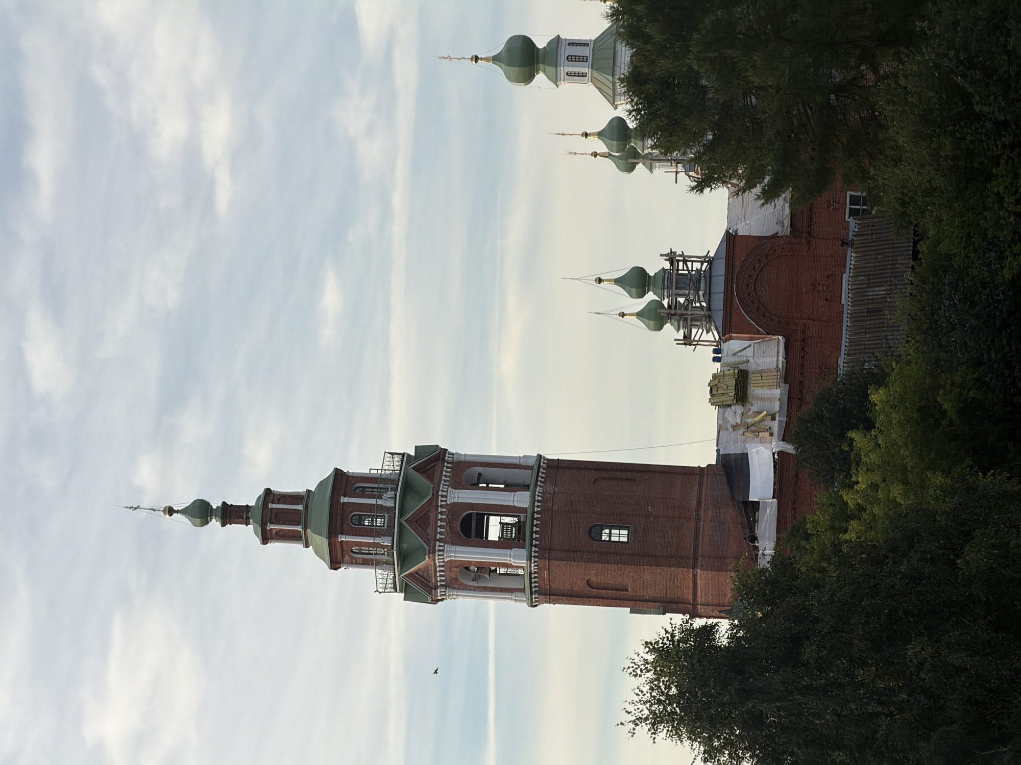 An exterior, eye-level, vertical shot captures the bell tower and domes of a brick church in Cherdyn, Russia, against a cloudy sky. The bell tower, a tall, cylindrical structure with red brick and green accents, features multiple levels with arched openings, decorative columns, and a pointed green dome topped with a cross. Several green-domed chapels or smaller towers are visible to the right of the bell tower, some appearing to be under construction with scaffolding. The lower portion of the scene is obscured by lush green trees, with glimpses of red brick rooftops and construction materials from the church buildings. A single bird is visible in the sky to the left of the bell tower.