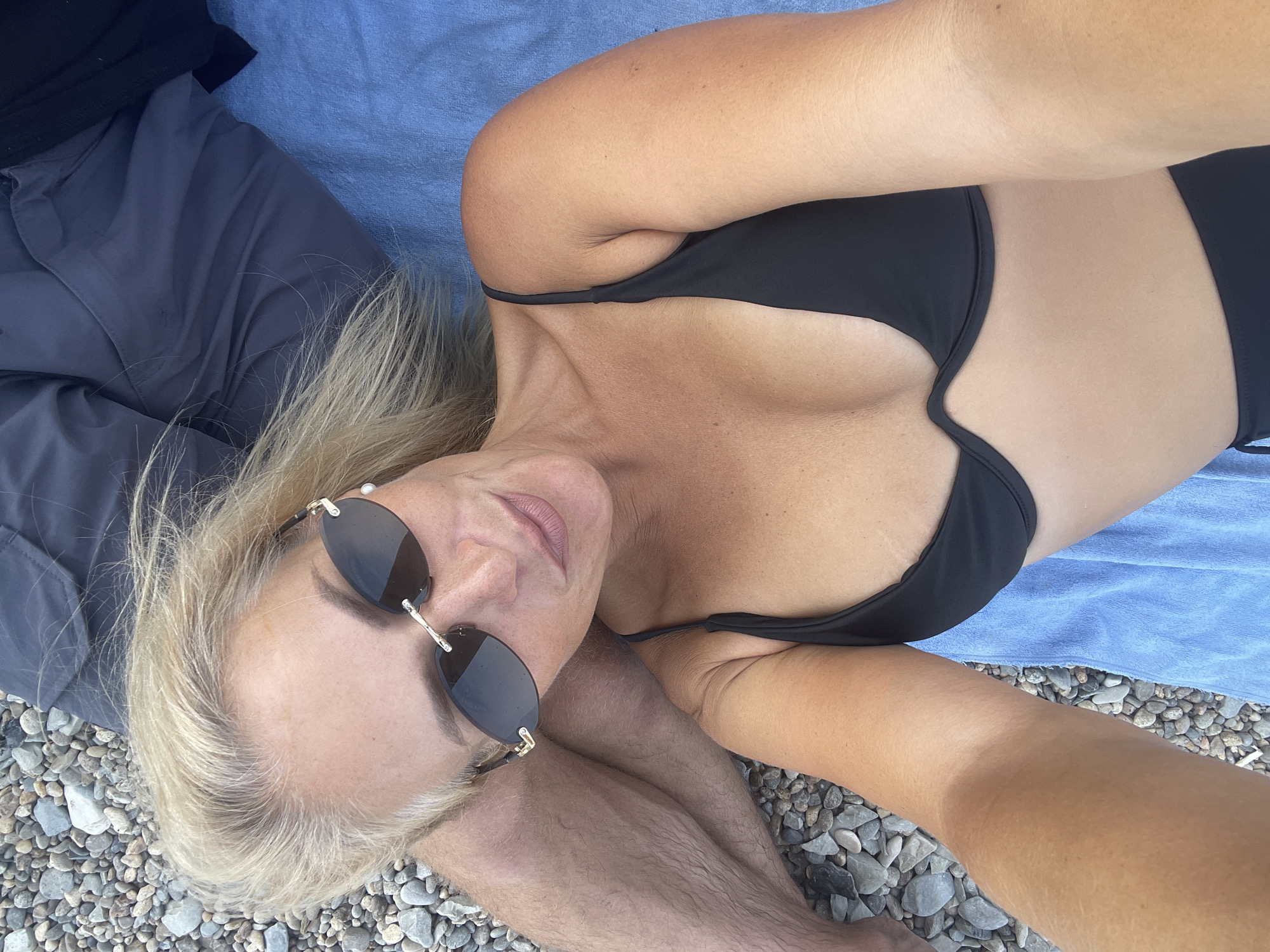 A woman is lying on a beach with gray pebbles. She is wearing a black bikini and sunglasses. Her hair is blonde and she is looking at the camera. A person is lying next to her, wearing gray shorts. A blue towel is under them. The scene is set in the city of Ol'ginskii, Russia.
