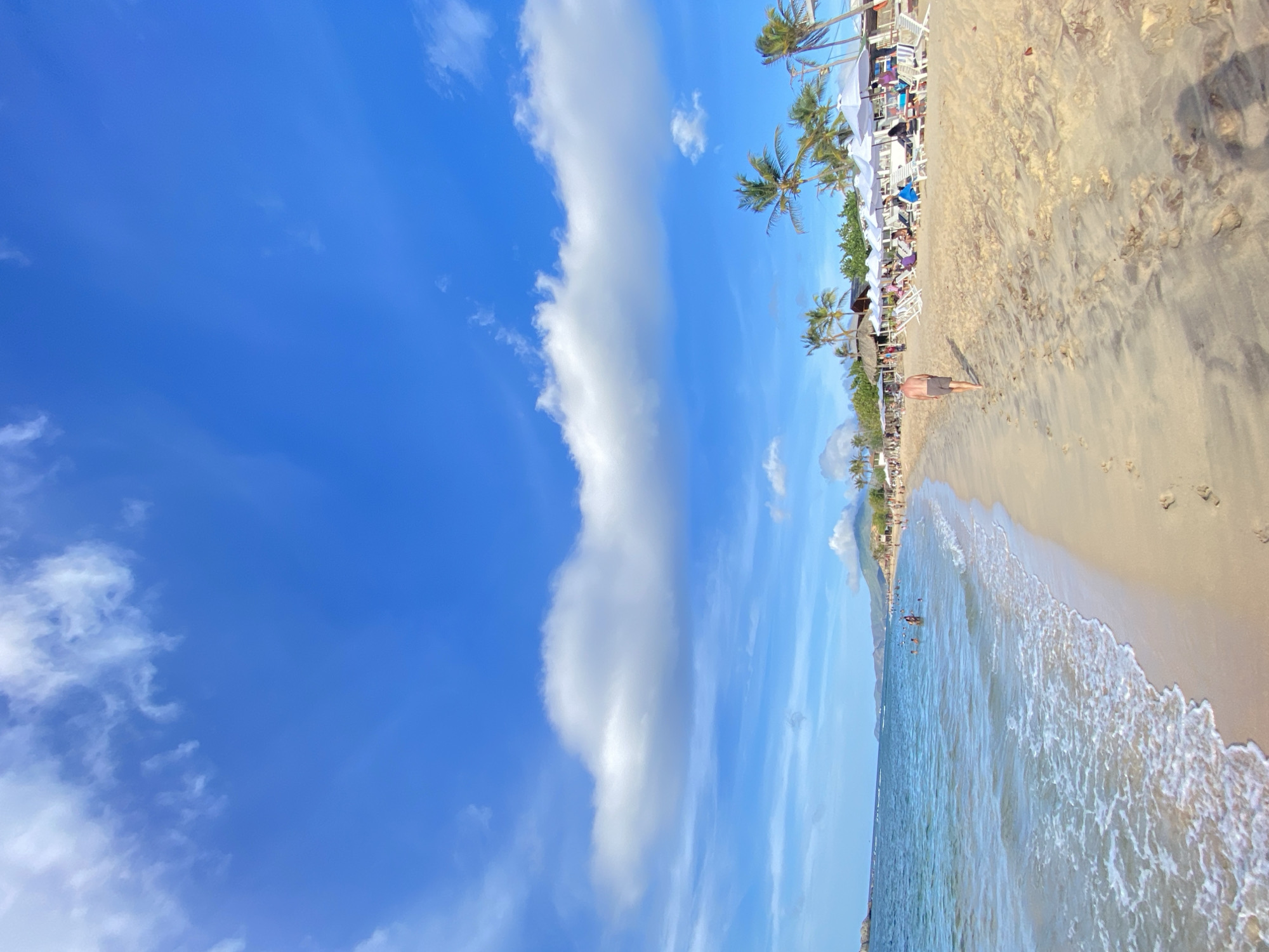 The image captures a sunny day at a crowded beach, likely in the city of Altagracia, Venezuela. The turquoise ocean meets a sandy shore lined with palm trees and white umbrellas providing shade for beachgoers. Several people are visible in the water and on the beach, some relaxing on lounge chairs and others walking along the shoreline. A lone man in shorts walks away from the camera down the sandy path. In the distance, mountains form a picturesque backdrop against the bright blue sky dotted with white clouds. The scene is indicative of a popular tourist destination, bustling with activity and enjoying the tropical weather.