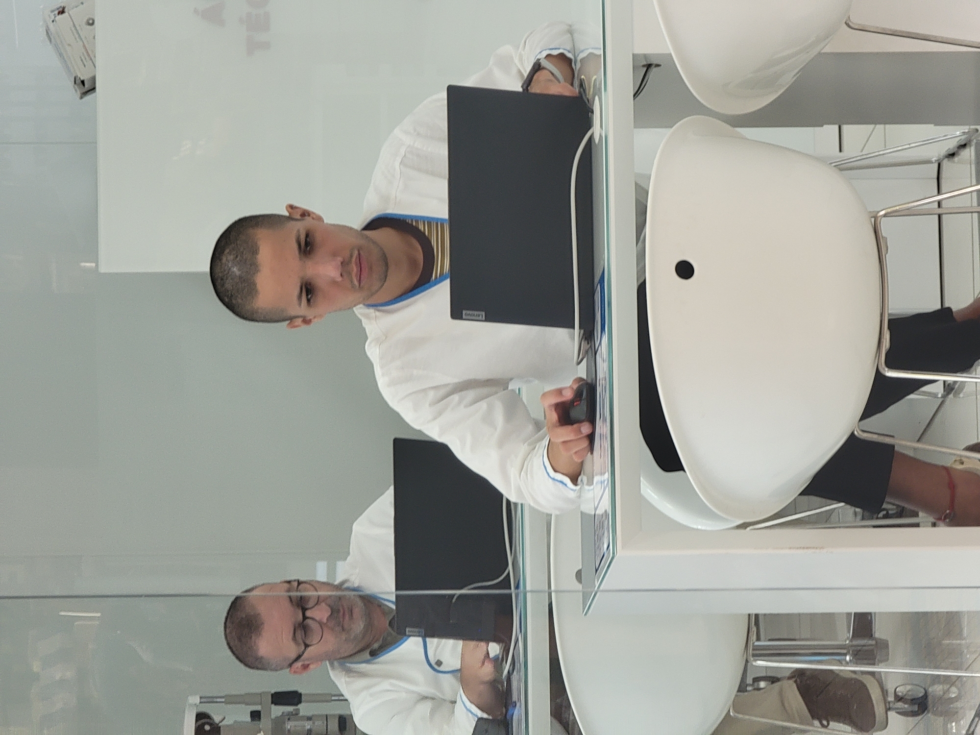 Two men are seated at a white table, each with a laptop. The man on the left, wearing glasses, is looking down at his laptop. The man on the right, with a shaved head, is looking at his laptop, with his hand on a mouse. Both men are wearing white shirts. The scene appears to be an office or classroom setting.
